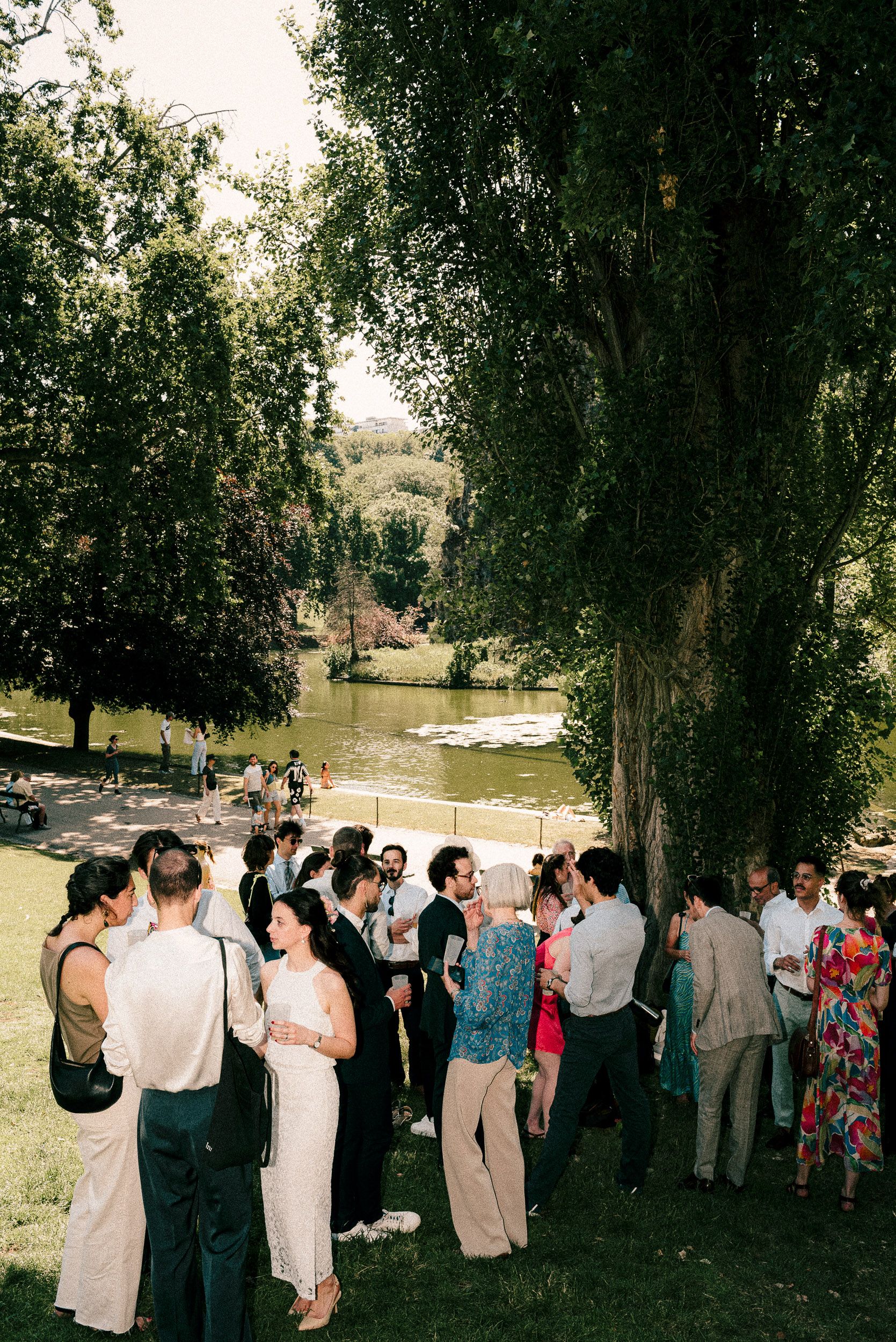 Wedding guests gather under shady trees near a lake during an outdoor celebration.