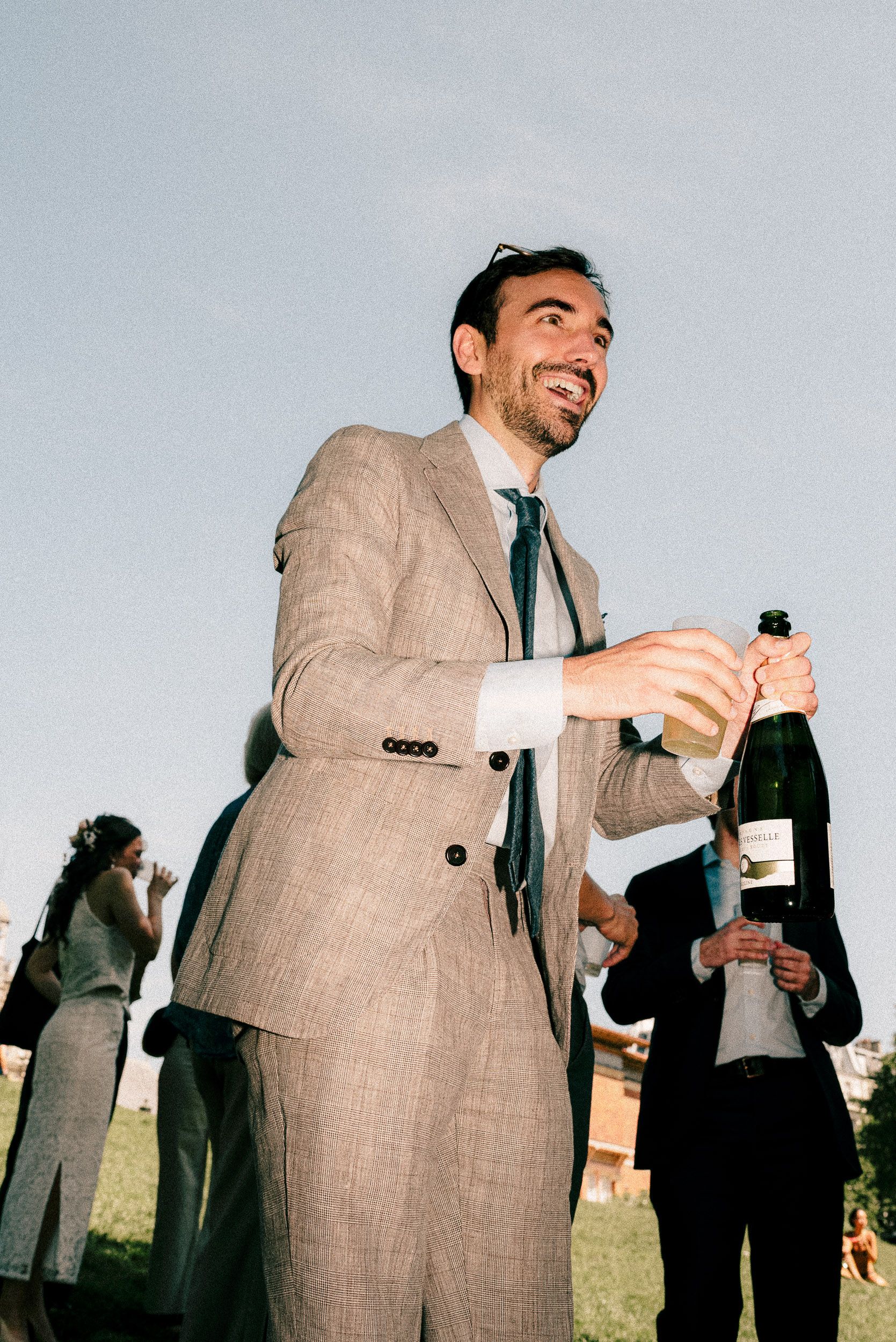 A person in a beige suit and tie smiles while holding a champagne bottle at an outdoor event.
