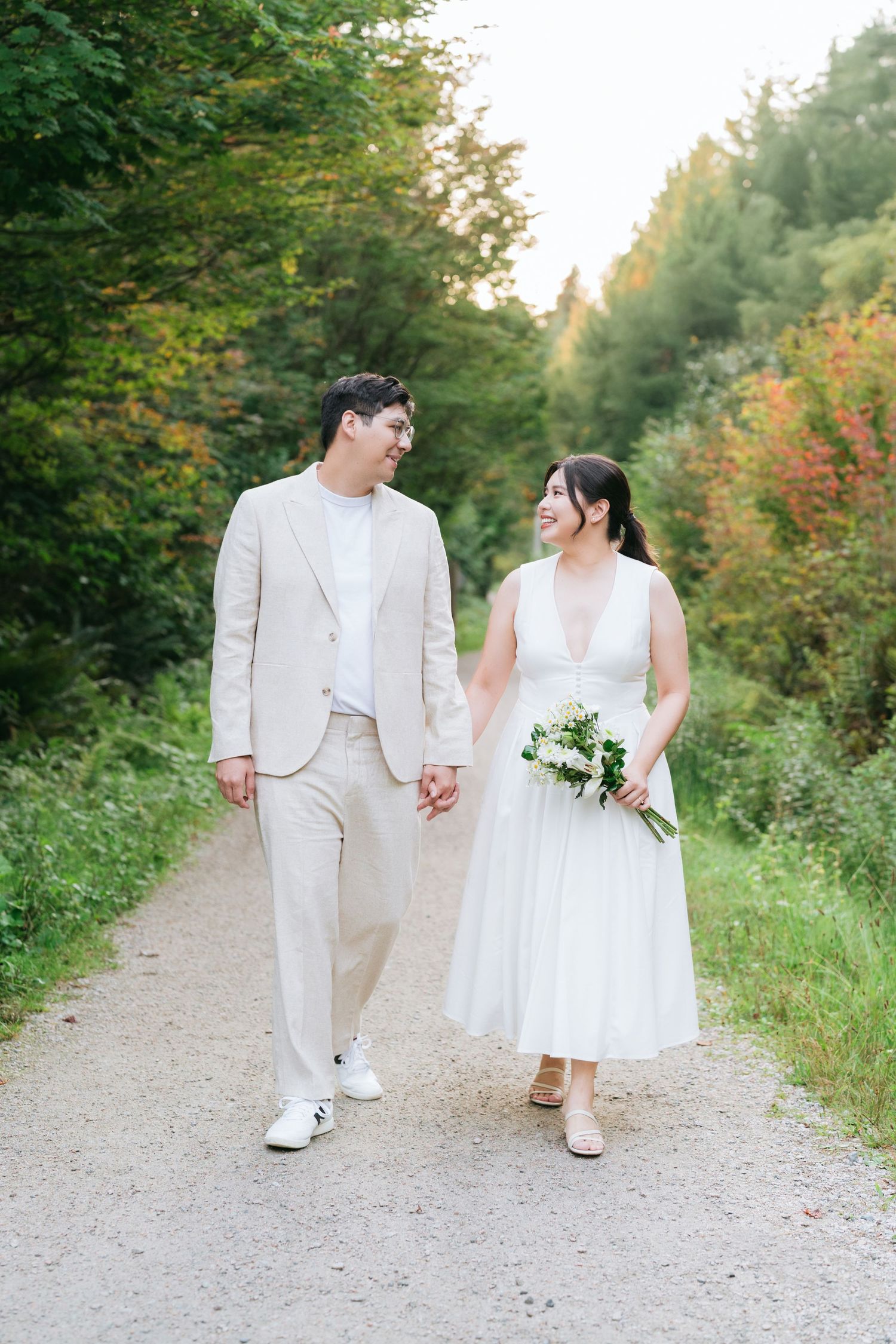 A couple in wedding attire walks hand in hand down a gravel path surrounded by lush summer greenery.