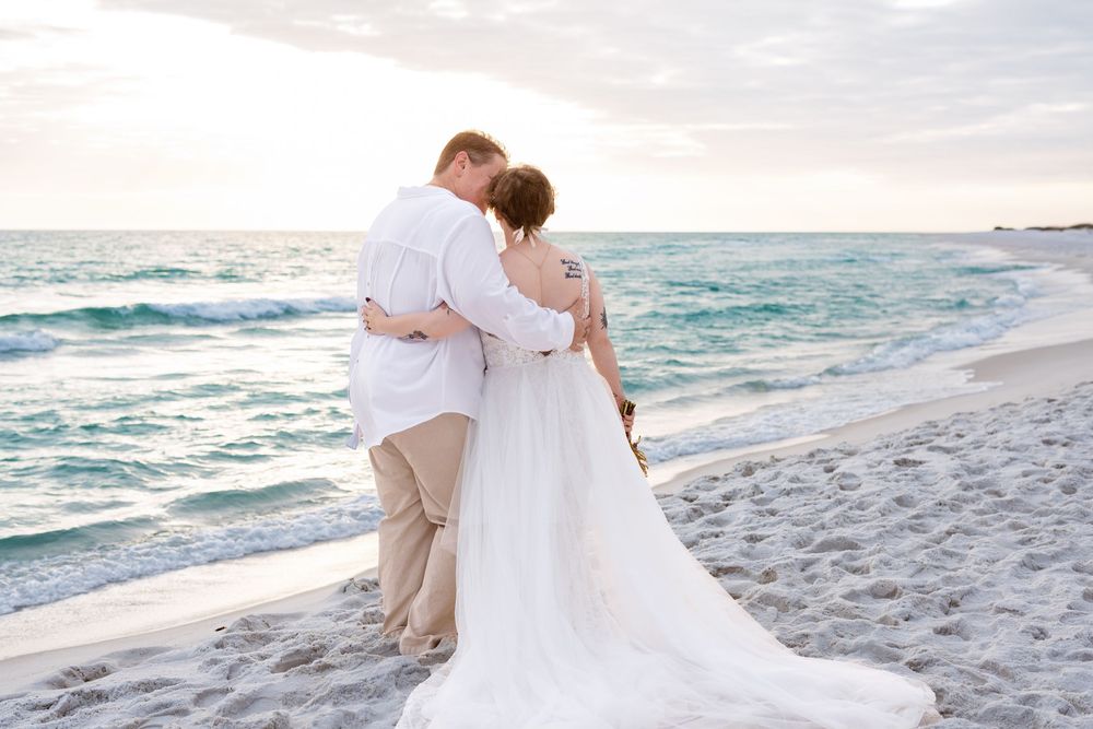A couple in wedding attire embraces while standing on a white sandy beach at sunset with turquoise waves rolling in.