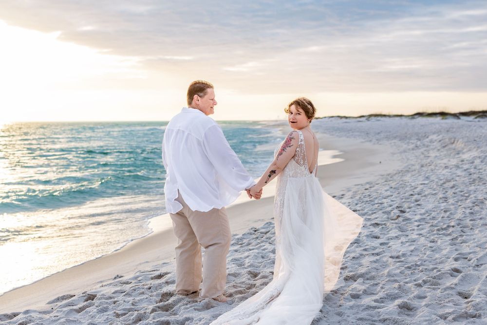 A couple in white attire walks along a pristine beach during sunset, creating romantic silhouettes against the ocean.