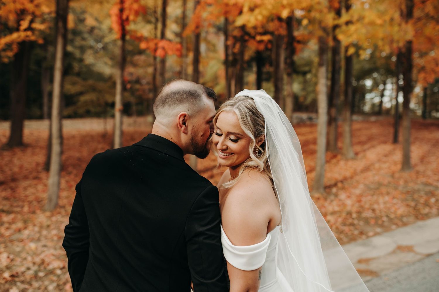 Bride and groom share a tender moment among vibrant autumn foliage, bride in white gown with veil.