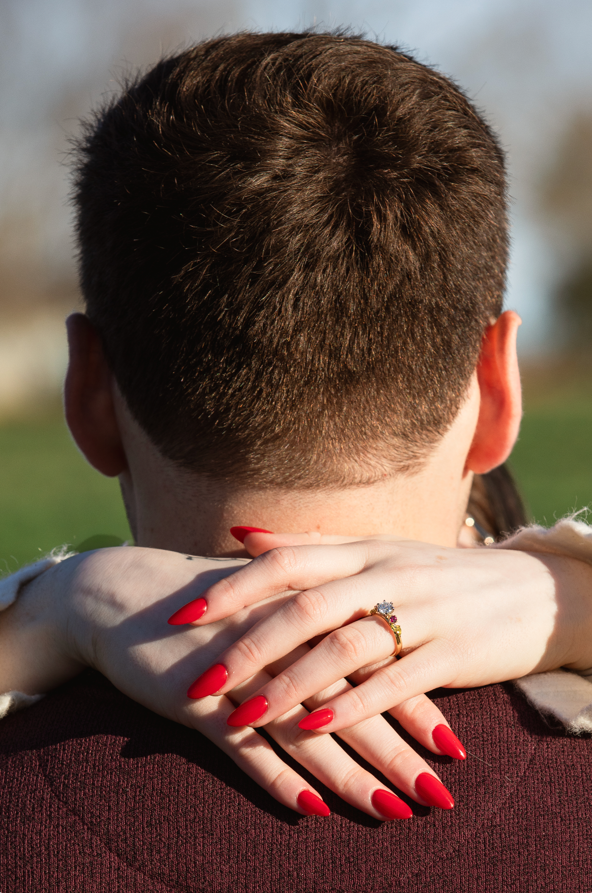 Woman with bright red manicured nails embraces someone wearing a burgundy sweater from behind showing off her engagement ring