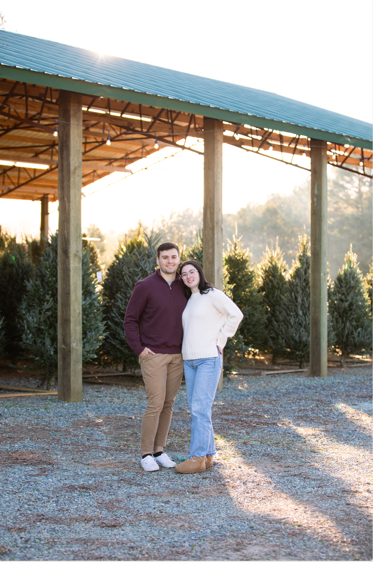 A couple poses together at a Christmas tree farm with evergreen trees in the background during golden hour.