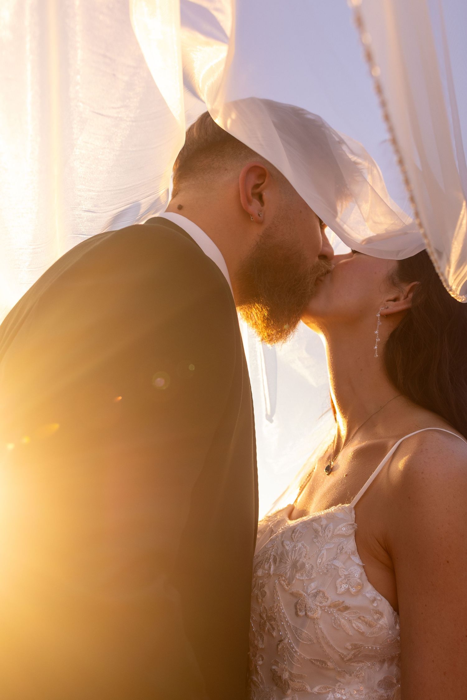 A romantic wedding couple shares a tender kiss at sunset with white curtains flowing in the background.