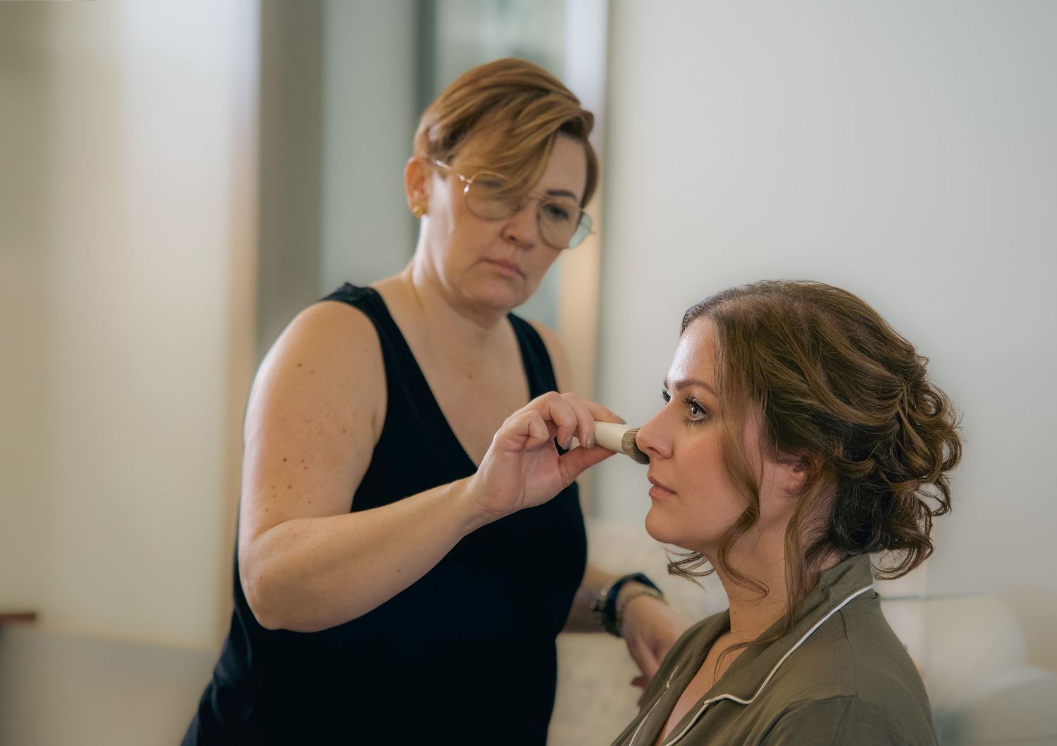 A makeup artist in a black tank top applies lipstick to a client with an elegant updo hairstyle.
