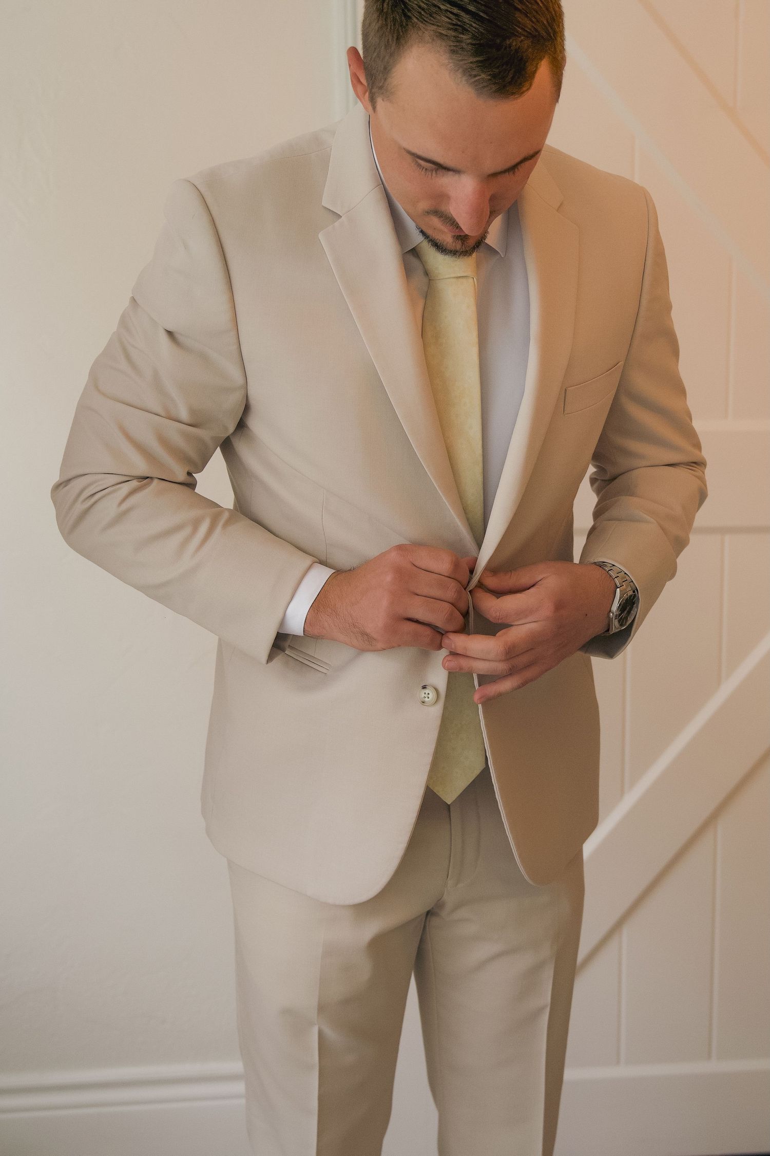 A person in a beige suit and yellow tie buttons their jacket while getting dressed for a formal occasion.