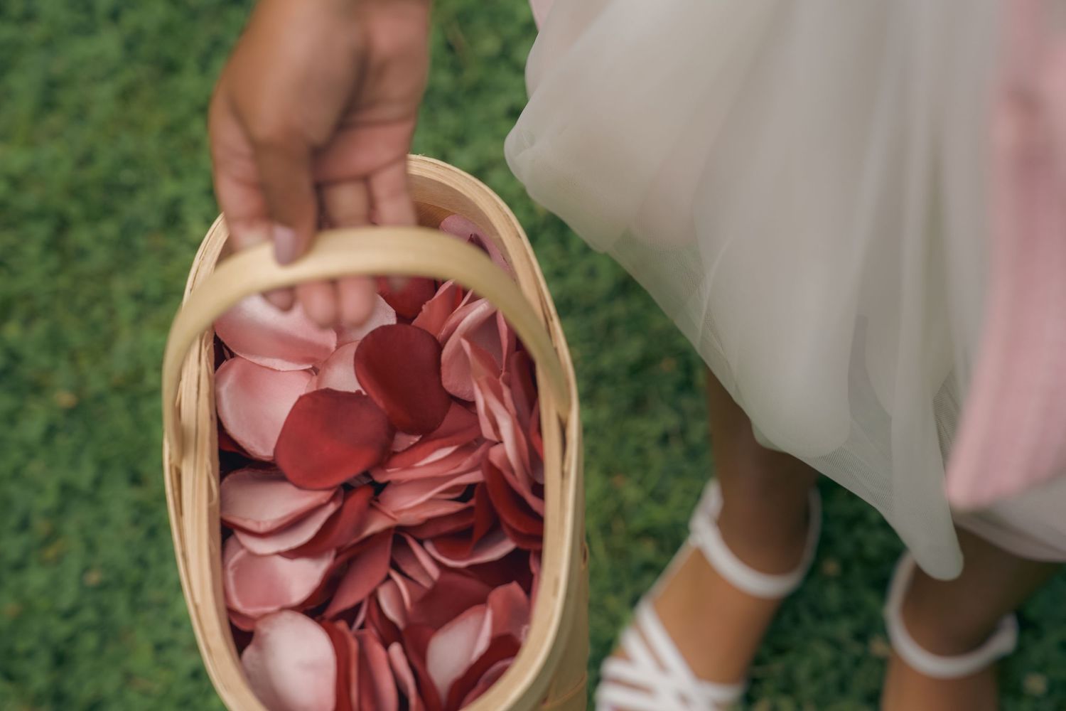 A woven basket filled with pink rose petals sits on green grass next to a person wearing white shoes.