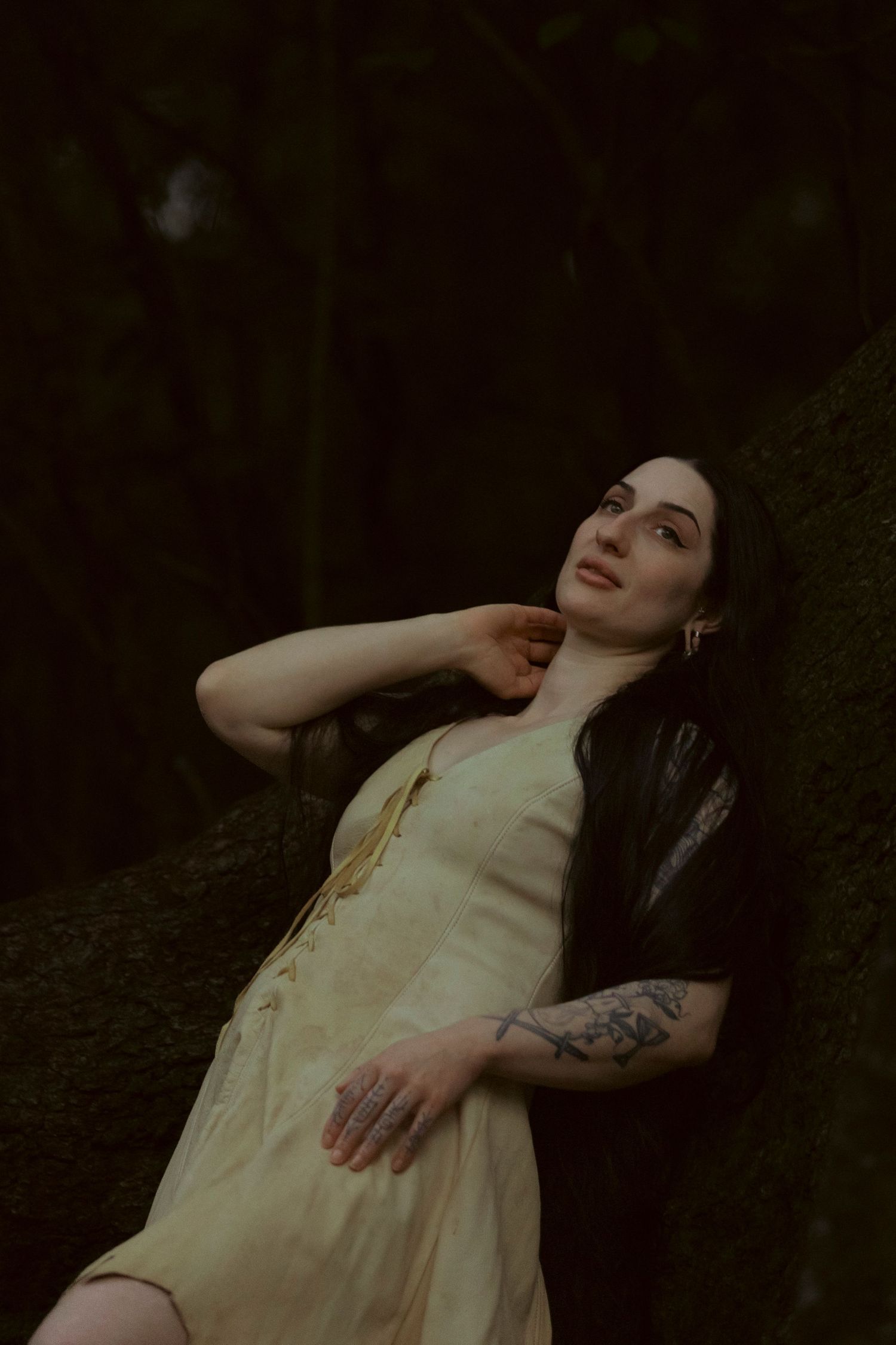 A figure in a light colored dress poses expressively against a dark forest backdrop in a moody artistic series.