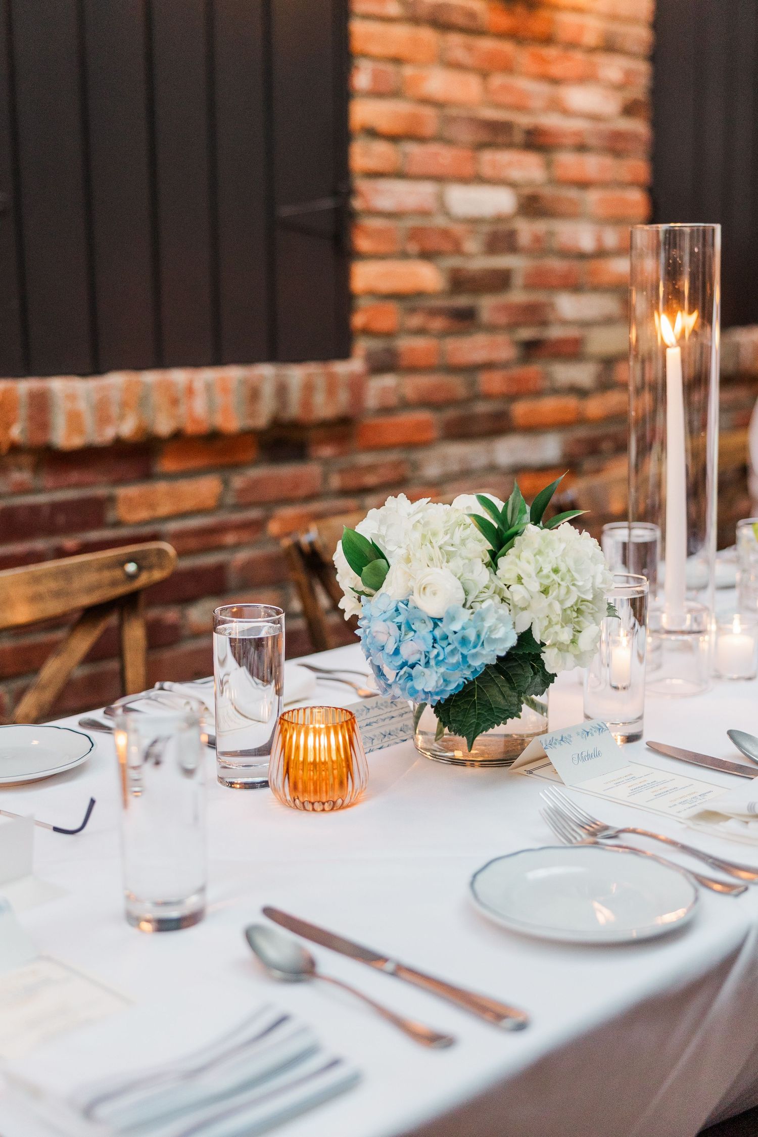 Elegant table setting with white linens, blue hydrangeas, and glowing orange candles against exposed brick wall.