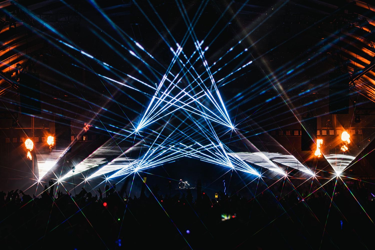 Stunning blue laser light show creates geometric star patterns at a concert with glowing orange torches on the sides.