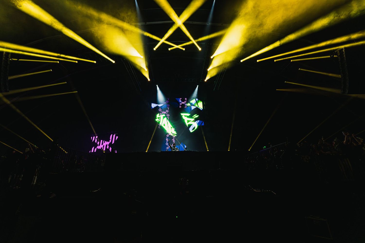 Concert stage illuminated with dramatic yellow spotlights and laser beams creating a dynamic lighting display during performance.