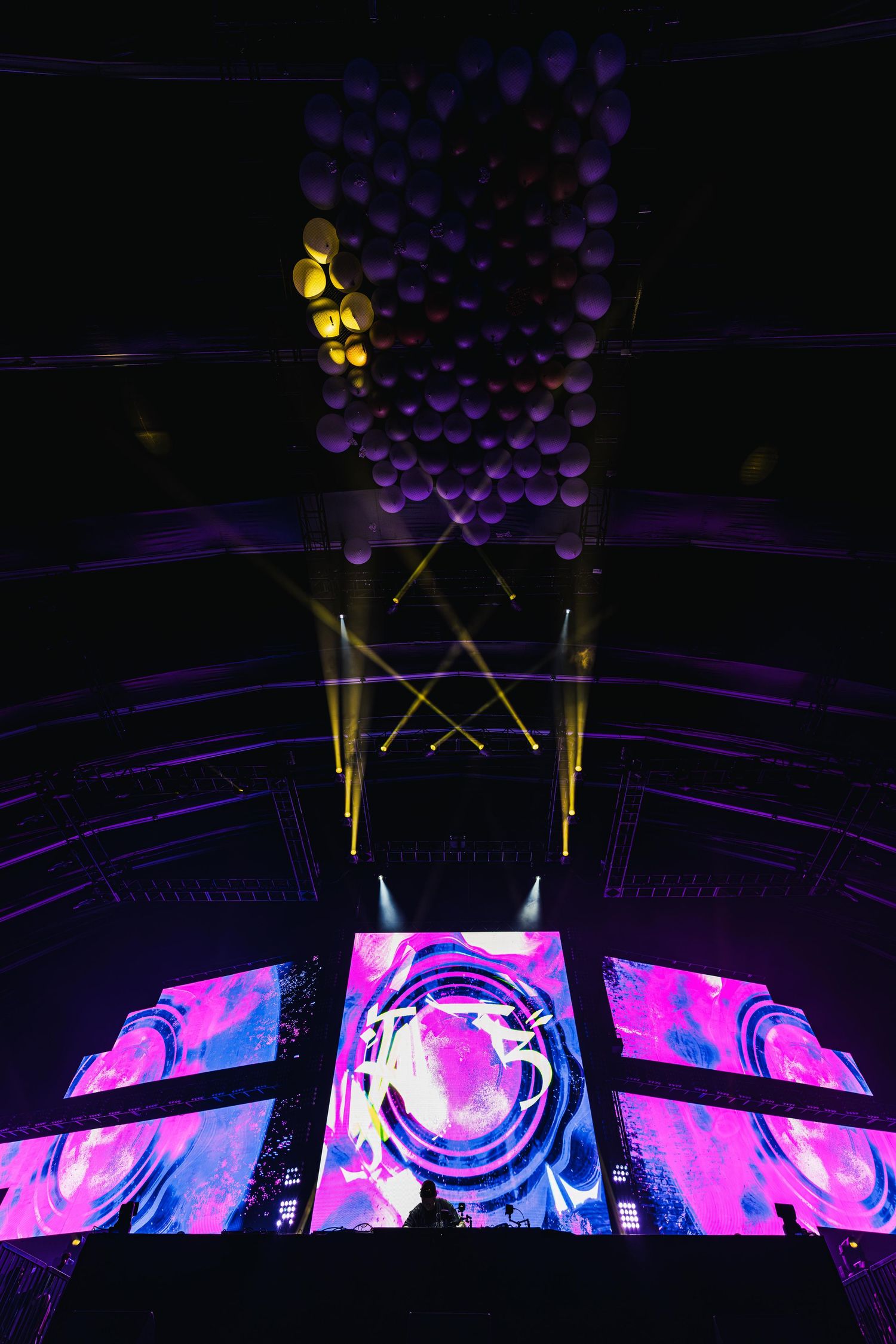 Concert stage setup with vibrant pink and blue LED screens displaying abstract patterns under a dark ceiling with suspended lights.