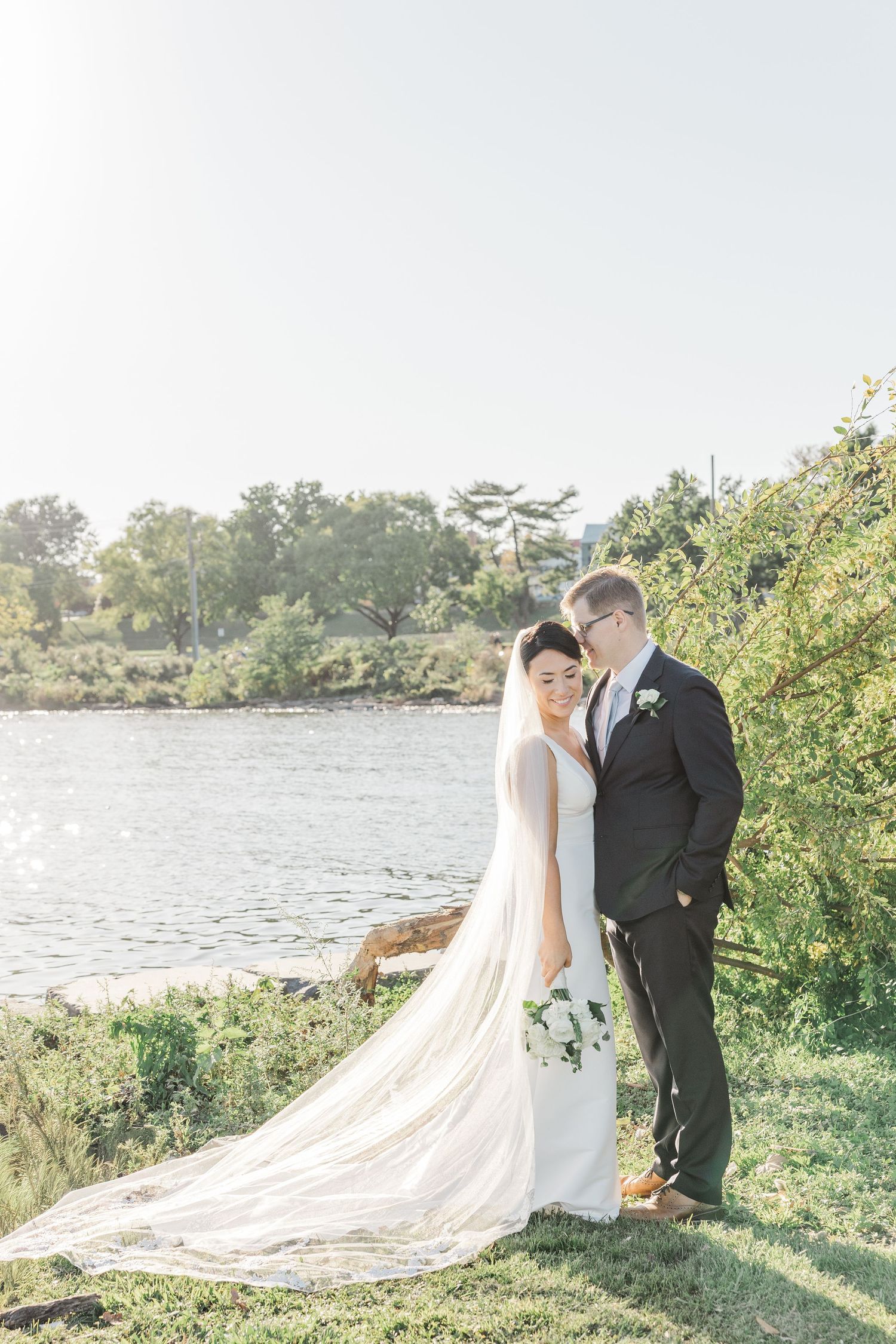 A romantic wedding couple stands by a serene lake with a flowing cathedral veil trailing behind them in the sunlight.