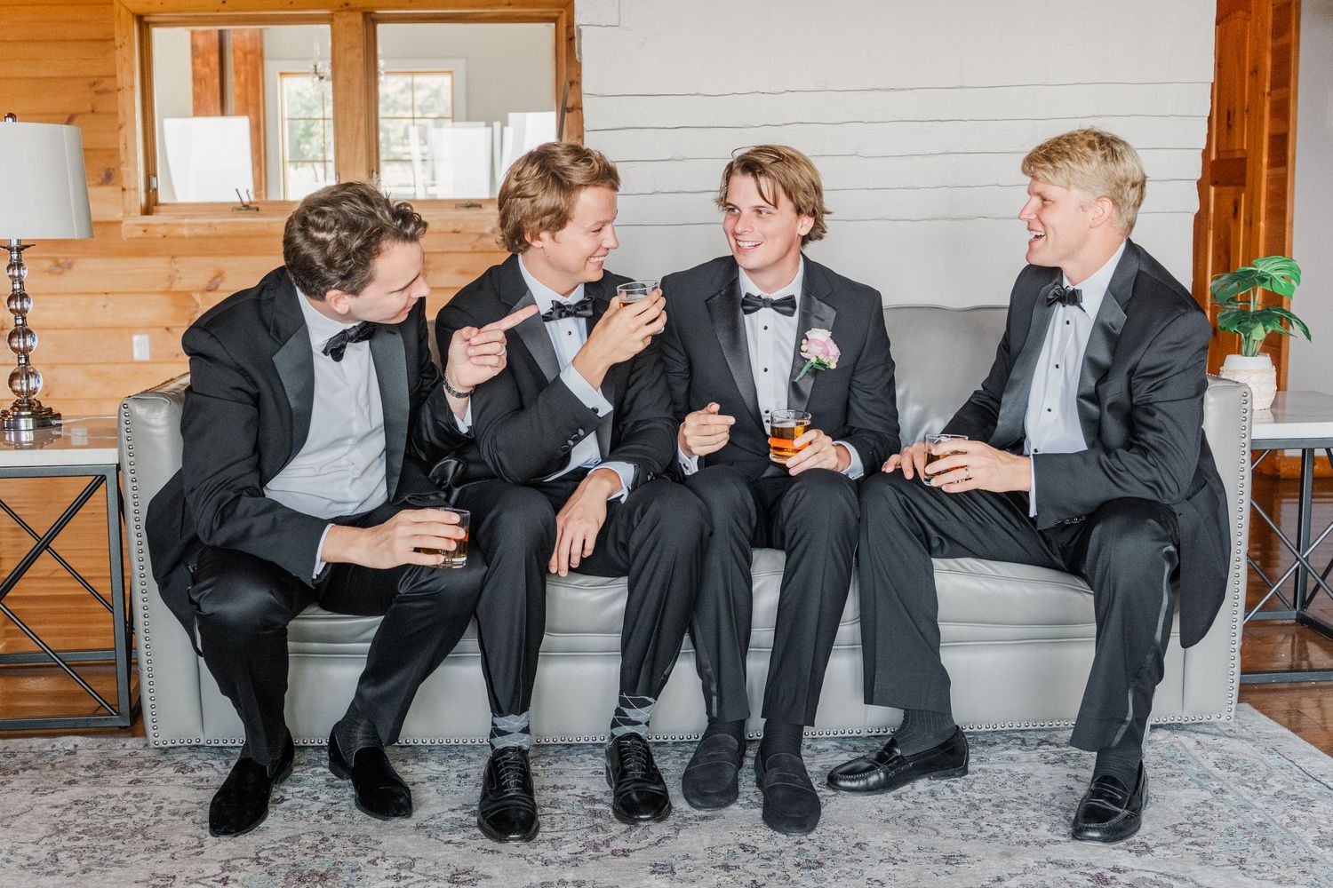 Groomsmen in black tuxedos sitting casually on white bench showing their dress shoes.