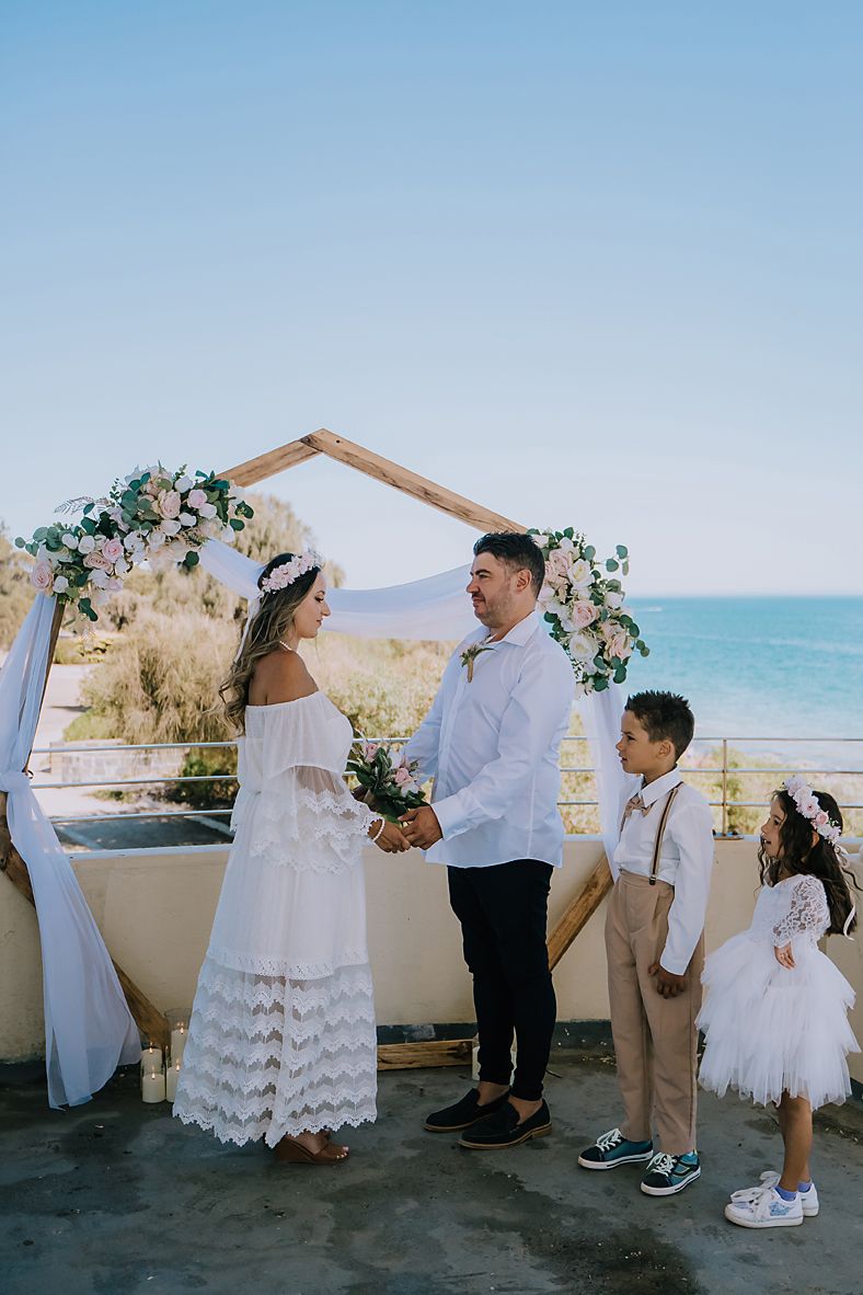 A beachside wedding ceremony under a wooden arch decorated with white flowers and fabric overlooking the ocean.