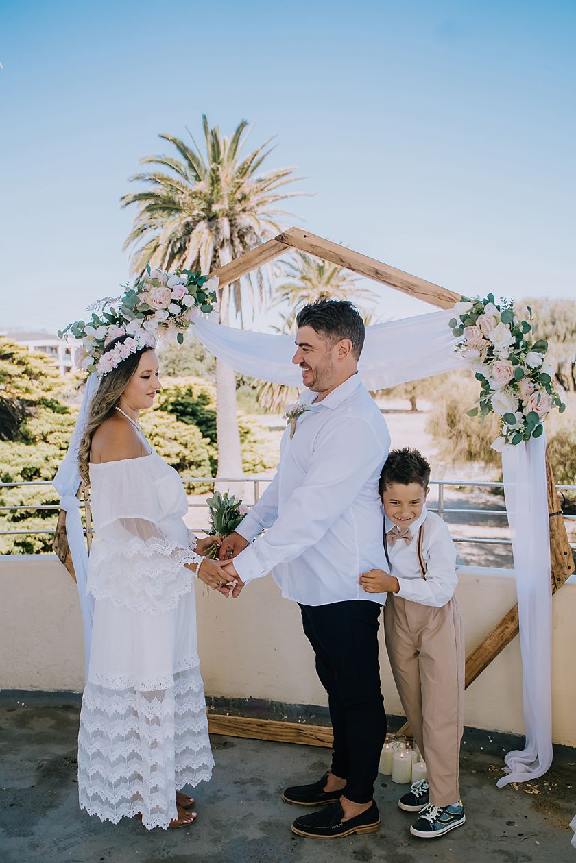 A romantic outdoor wedding ceremony with palm trees and a white floral arch in a desert setting.