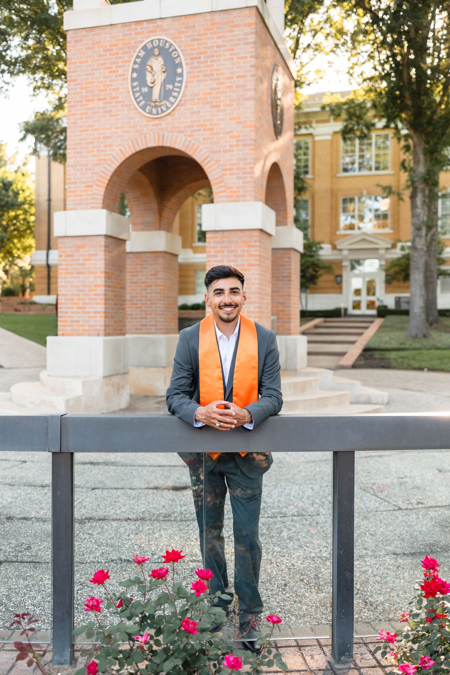 Smiling male graduate in orange stole poses at Sam Houston State University brick arch entrance with roses.