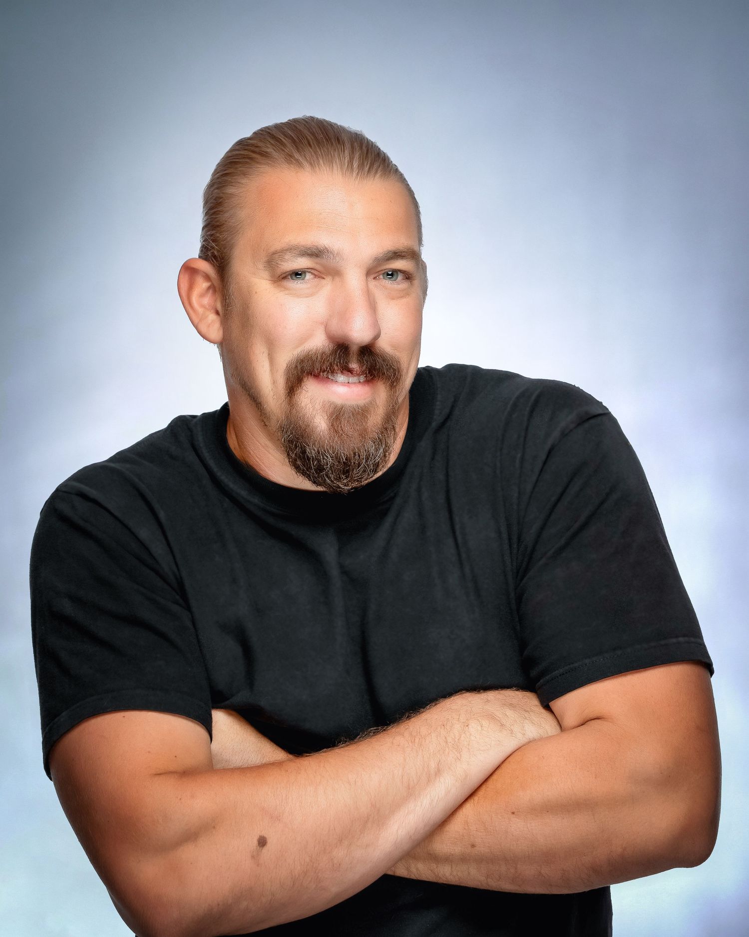 A confident man with blonde hair and beard poses with crossed arms in a black t-shirt.