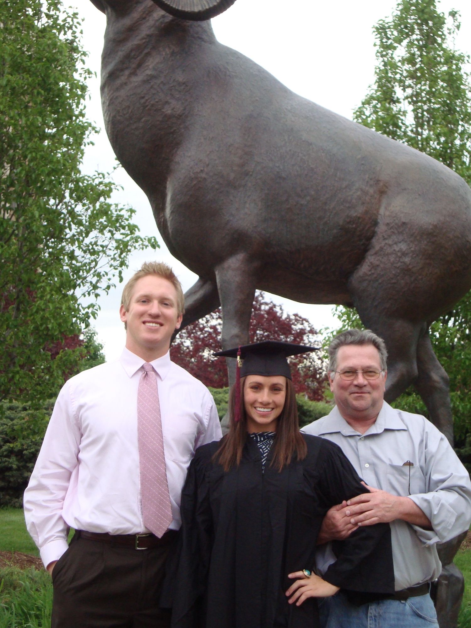 Three people stand next to a large bronze ram statue in a garden setting.