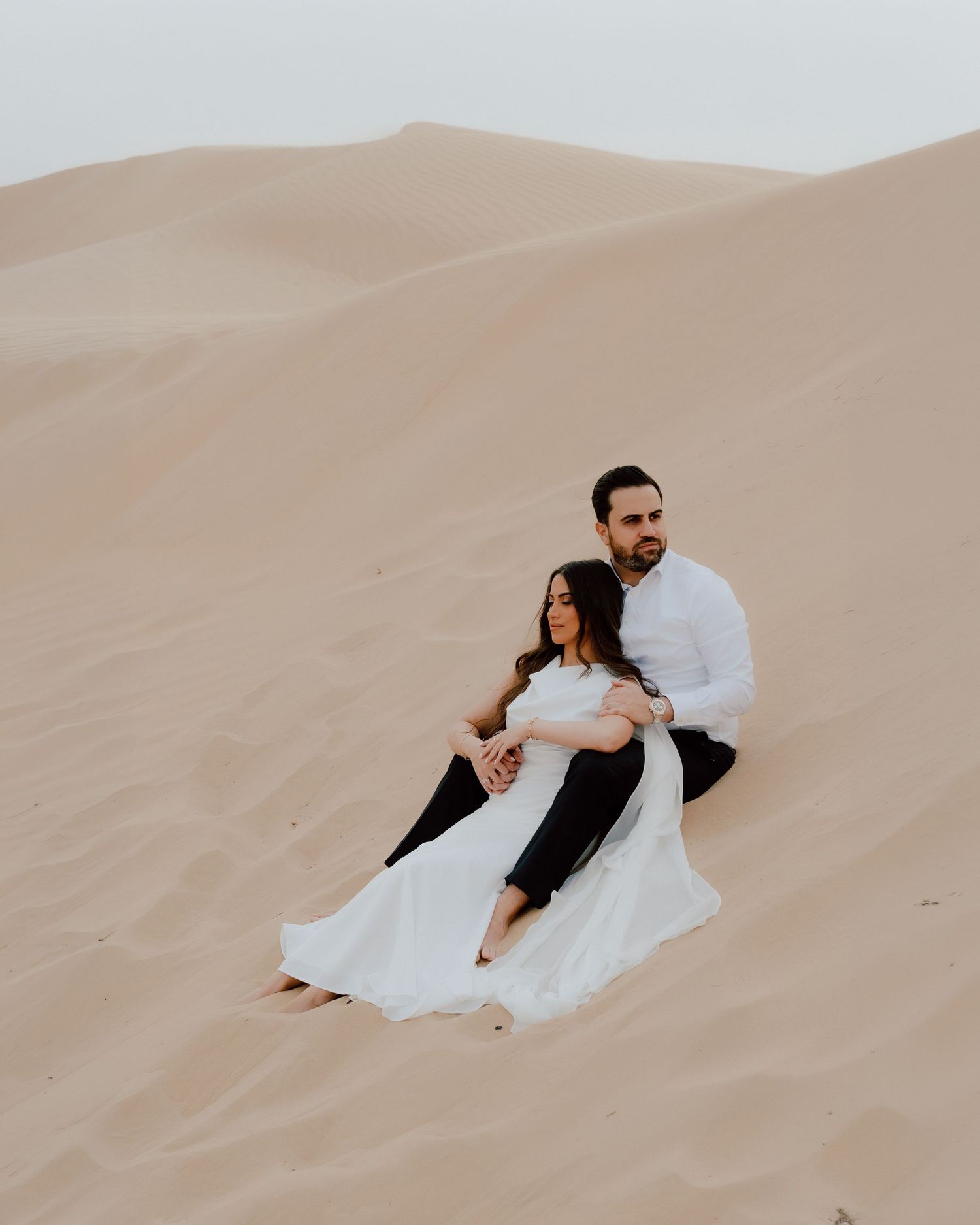 Couple sitting together on golden sand dunes in the desert, woman in white dress, man in white shirt and black pants.