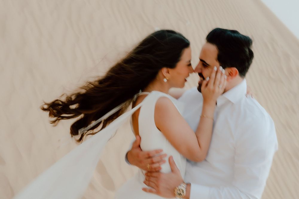 Romantic couple in white outfits nearly kissing on sandy desert dunes, woman's hair blowing in the wind.