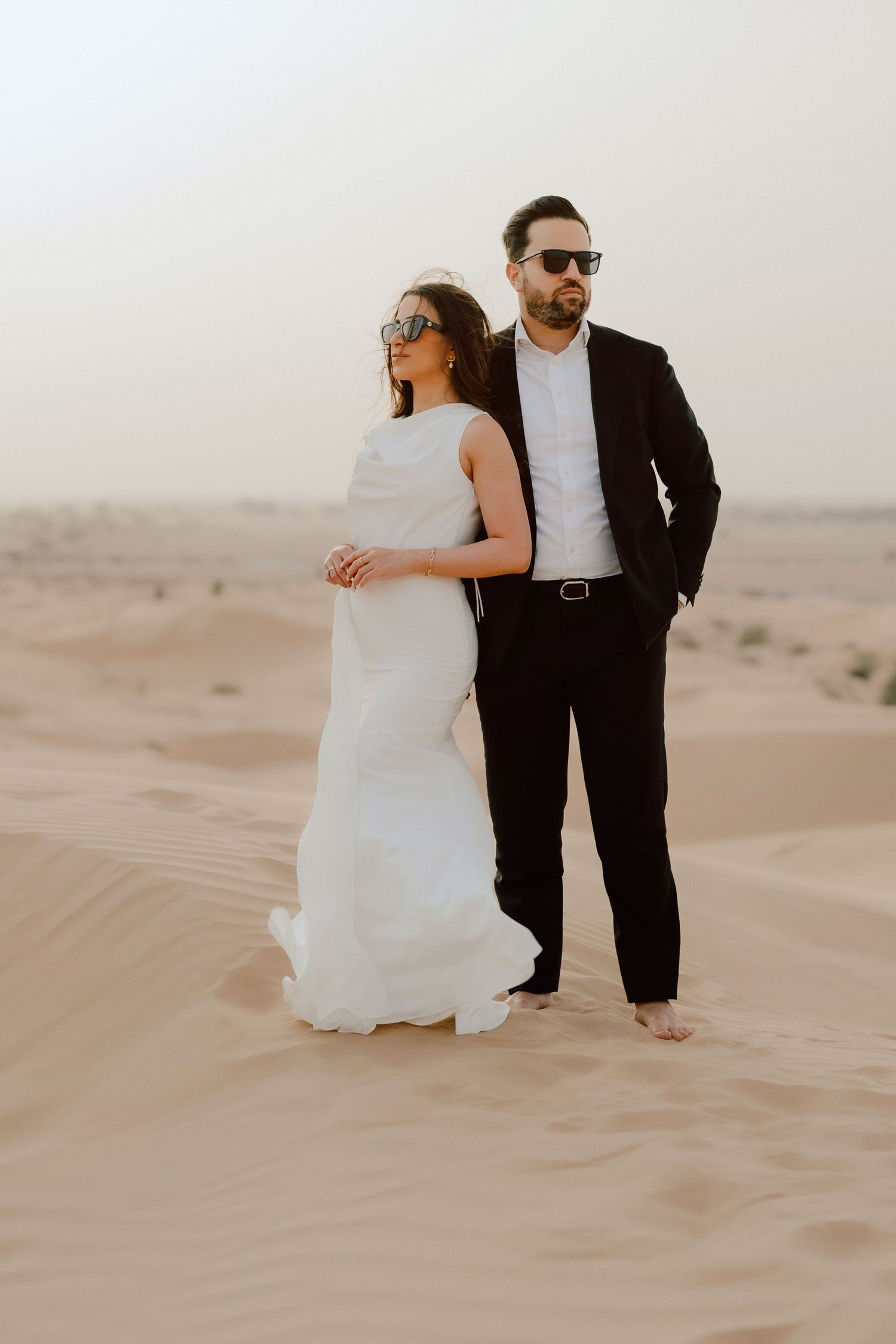 Couple in wedding attire posing on desert sand dunes, bride in white gown and groom in black suit wearing sunglasses.