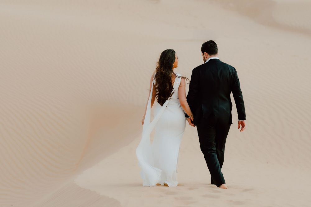Bride and groom walking hand in hand across sandy desert dunes, bride in flowing white gown, groom in black suit.