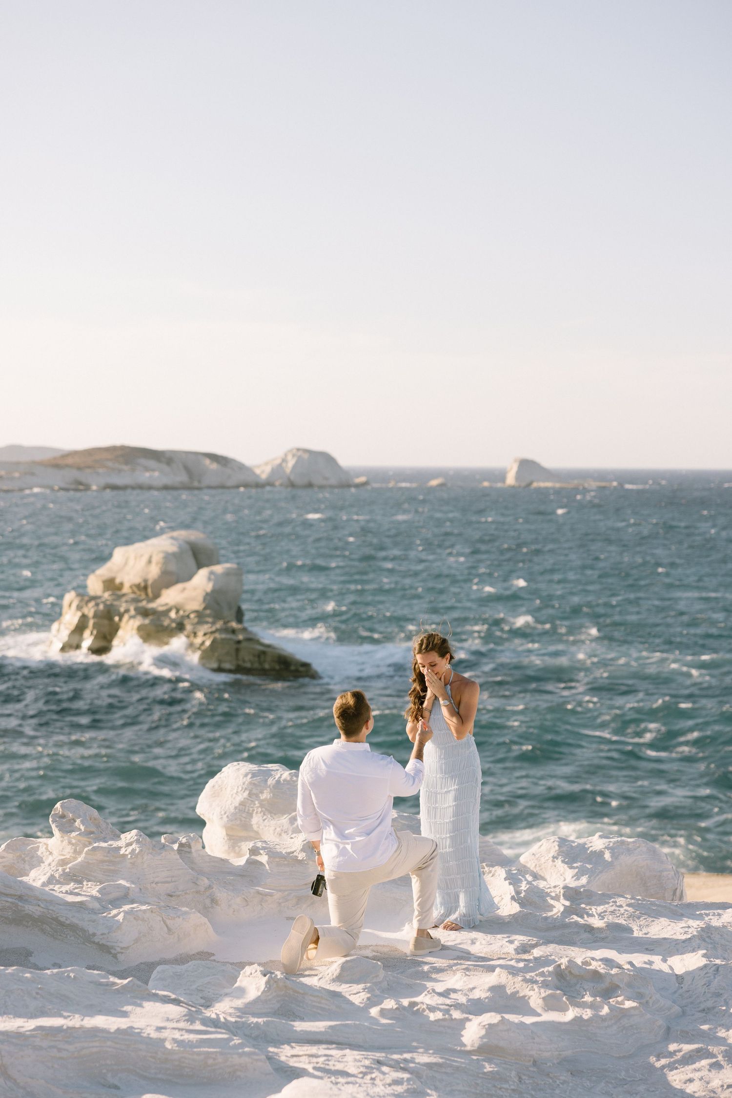 Couple in white clothing standing on snow-covered rocks by the ocean taking photos at sunset.