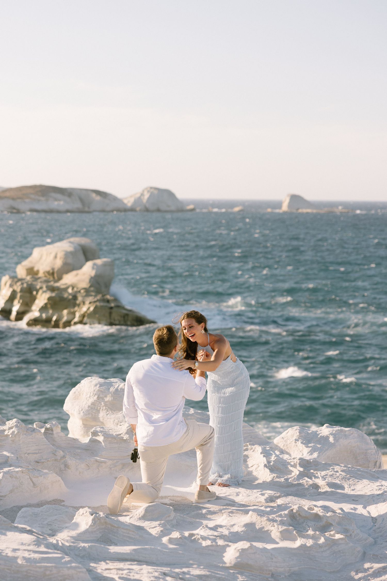 A couple in white clothing sits on snowy rocks by the ocean with white waves and cliffs in the background.