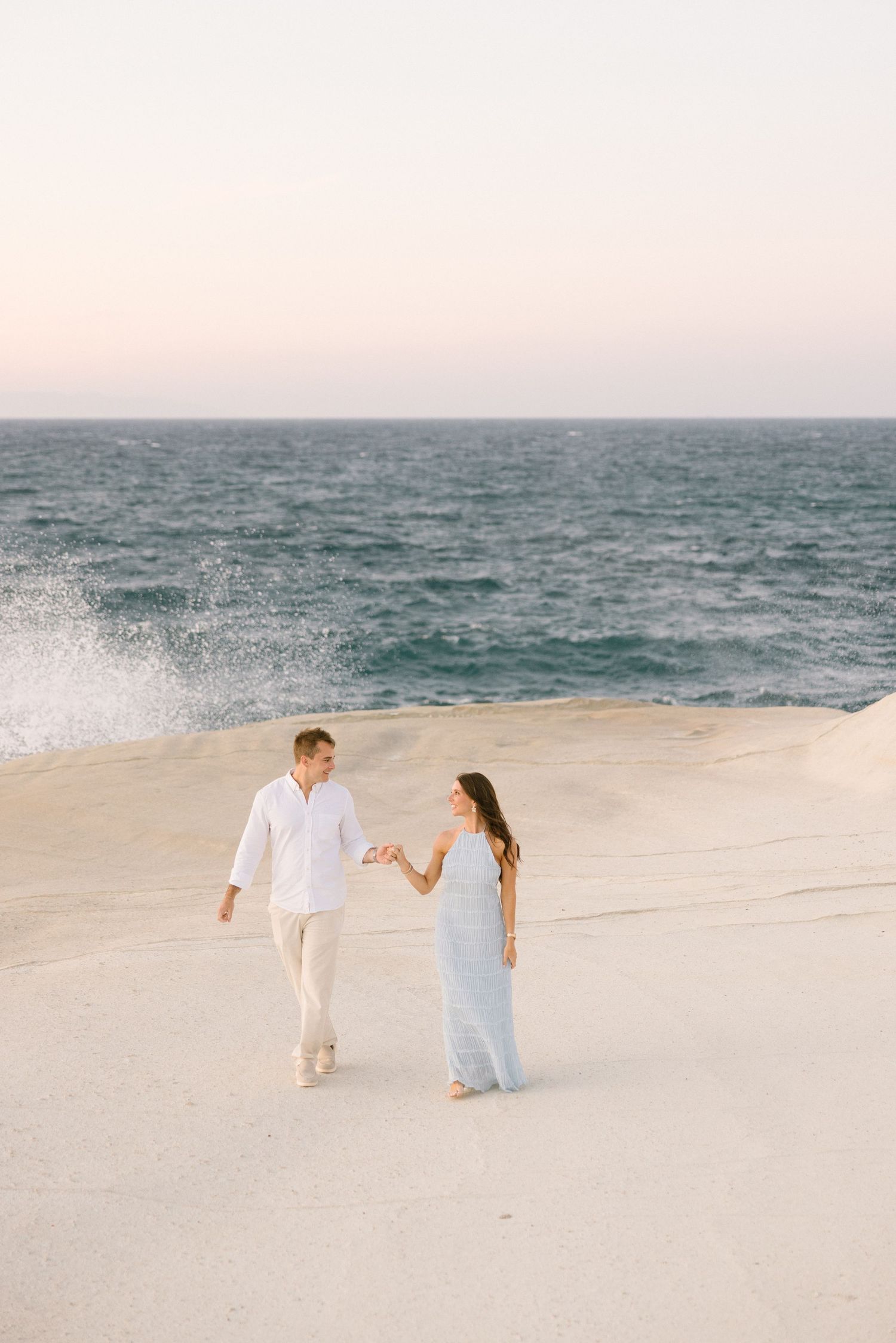 Couple walks hand in hand along a pristine white sand beach with ocean waves crashing behind them.