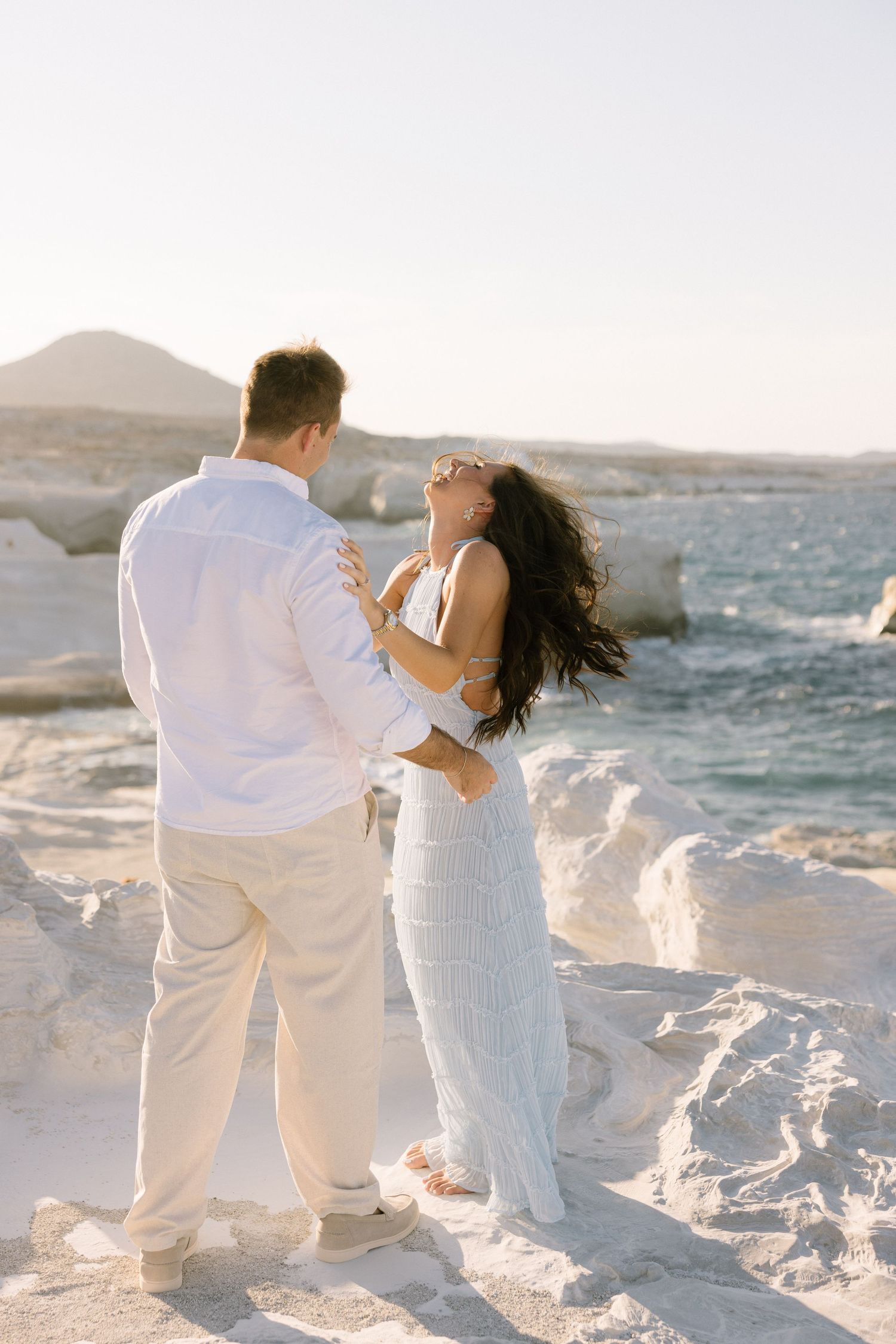 A romantic couple in white attire embraces on snowy coastal rocks at sunset.