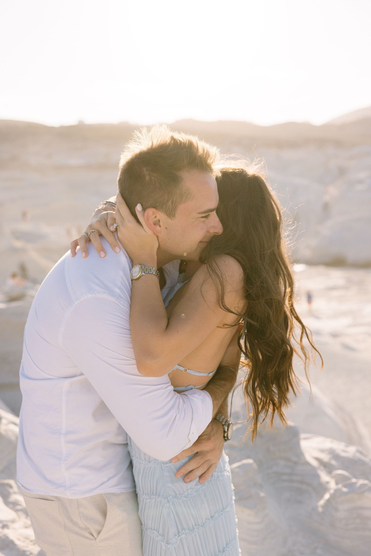 A couple shares an intimate moment on a beach during golden hour with desert hills behind them.