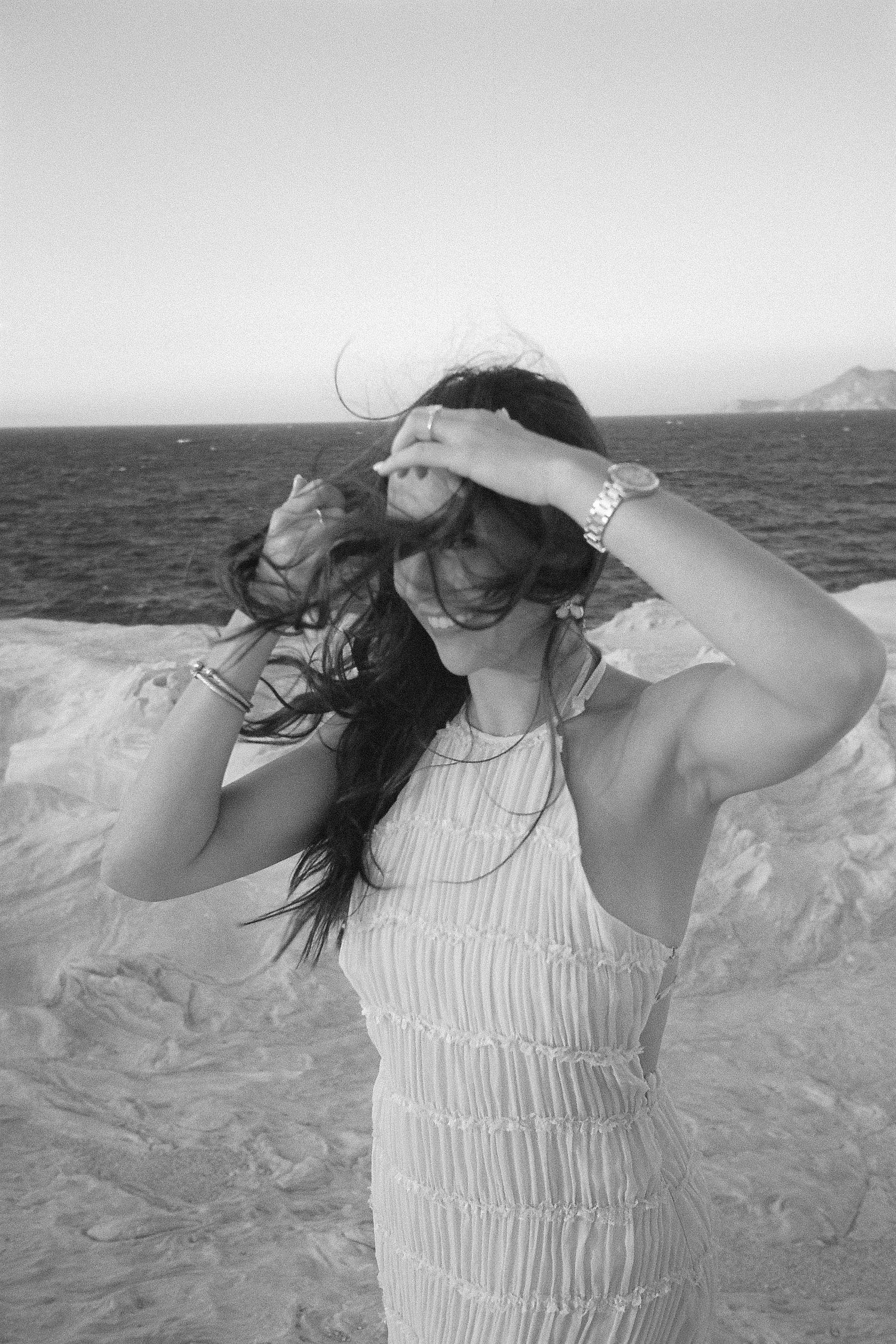 Black and white photo of someone in a textured white dress shielding their eyes from wind at a coastal location.