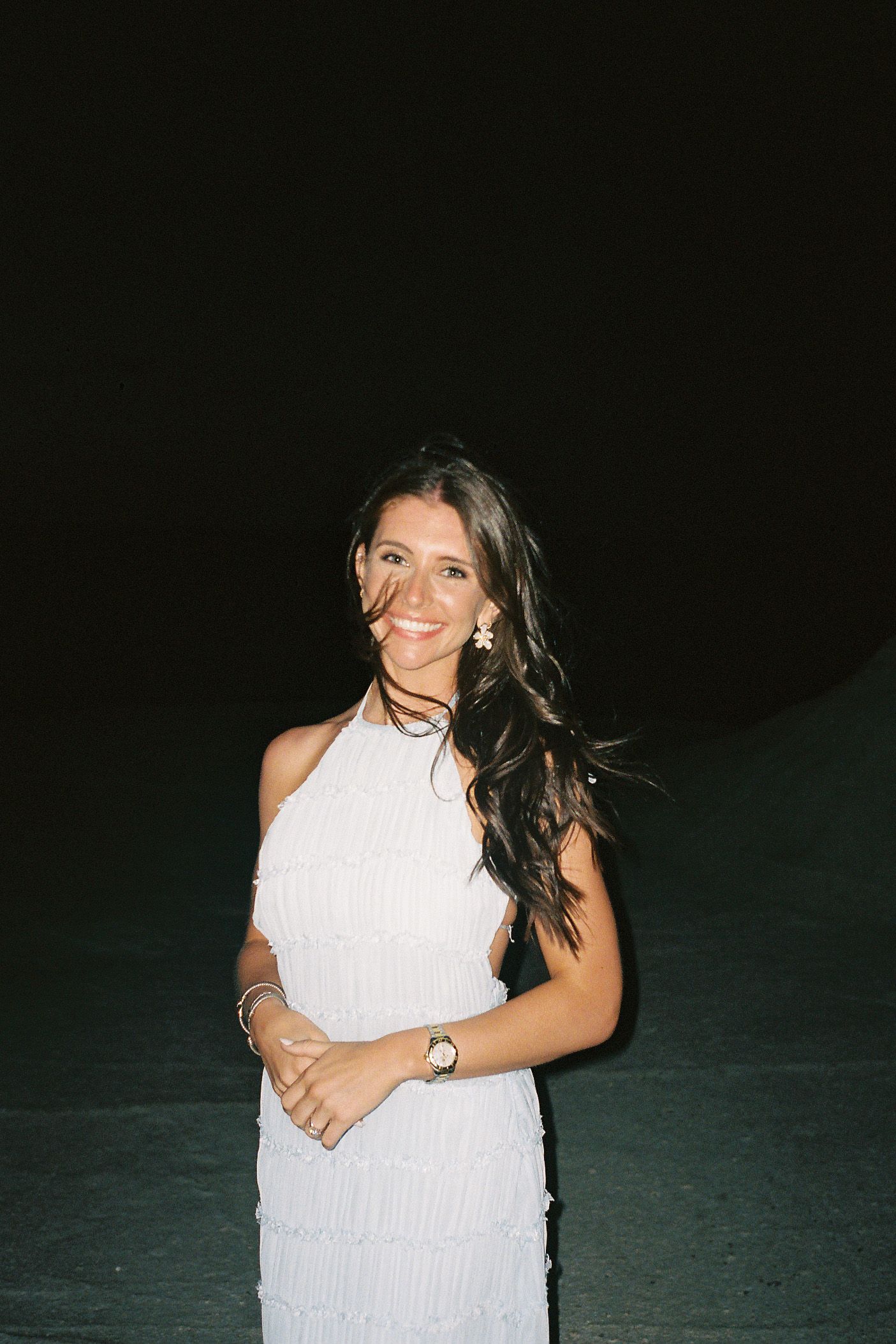 A person in a white sleeveless top smiles while posing against a dark background at night.