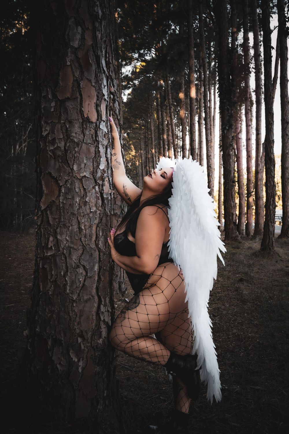 A figure wearing white angel wings poses artistically in a dark forest setting with tall pine trees in the background.