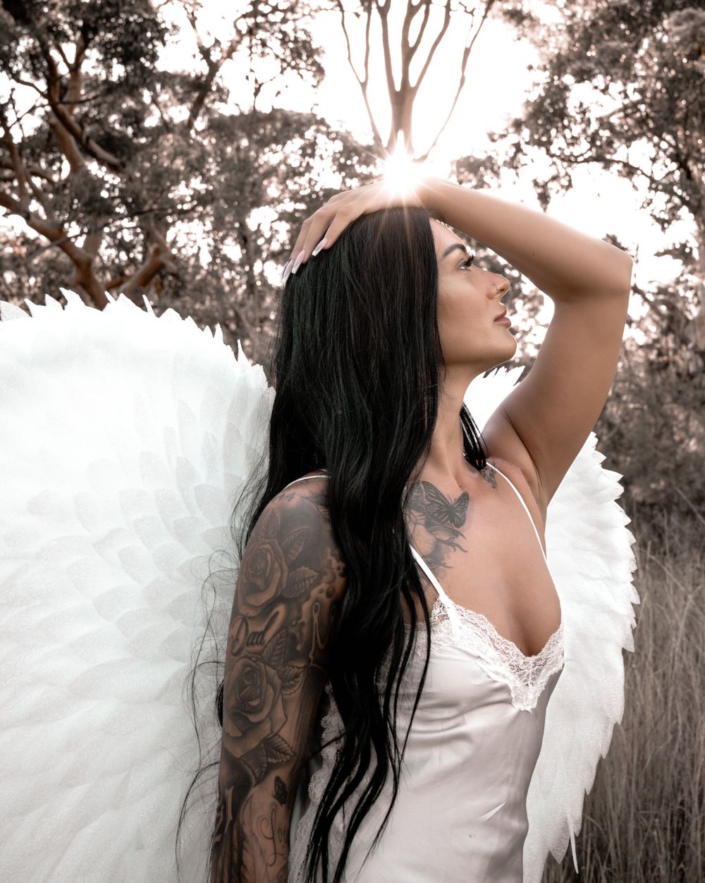 Woman with long dark hair and tattoos poses in white top against forest backdrop in artistic photo series.