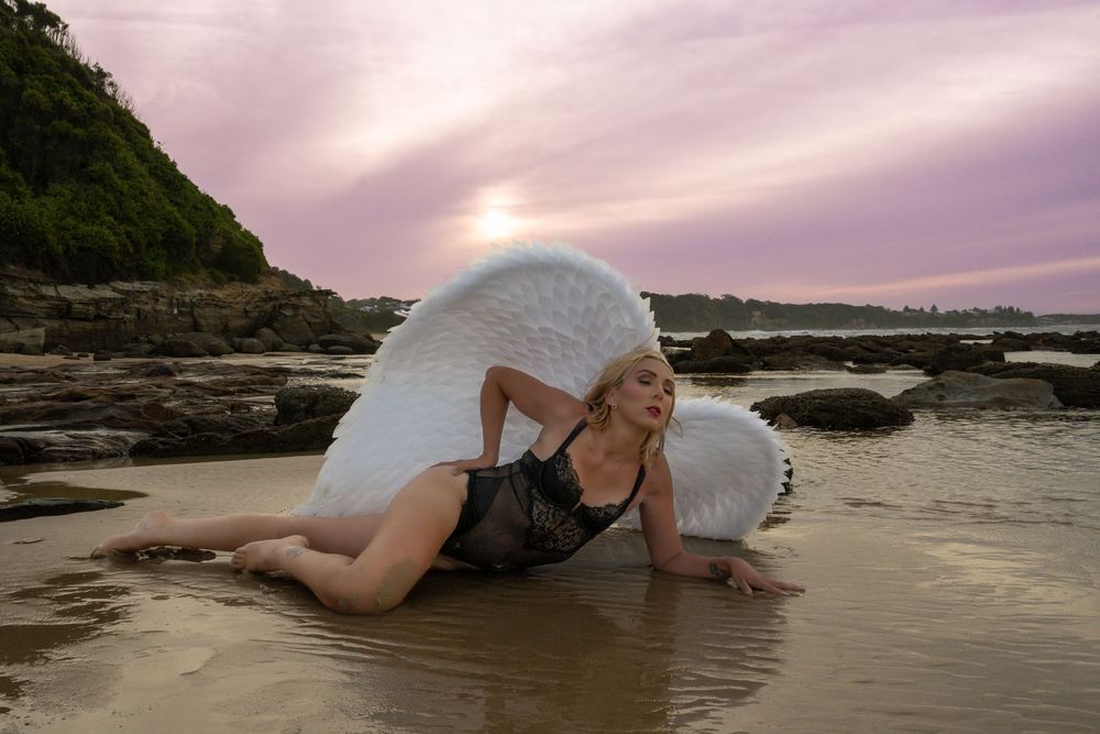 Woman in black swimsuit with angel wings poses on sandy beach at sunset with rocky coastline backdrop.