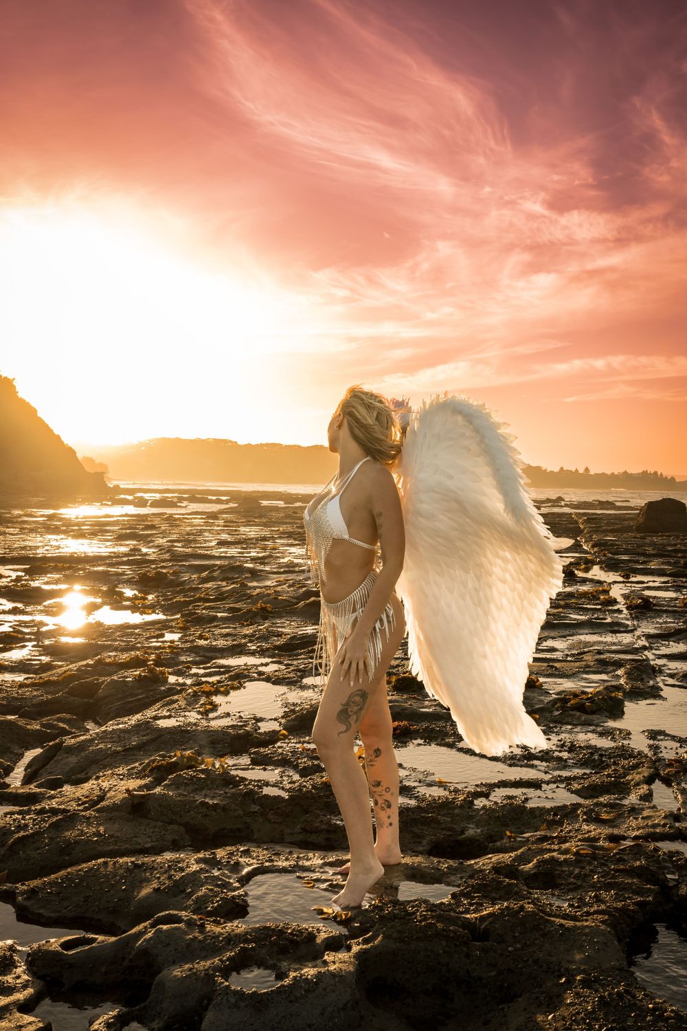 Blonde woman with large white angel wings stands on rocky shoreline at vivid sunset.