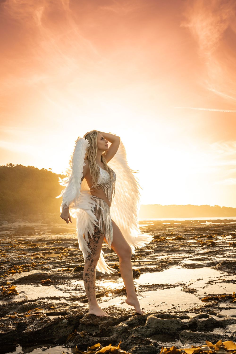 A woman in a white angel costume with feathered wings poses on rocky shore at golden sunset.