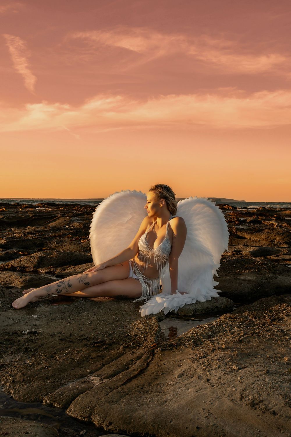 Woman in white angel wings and lingerie sits on rocky beach at golden sunset.