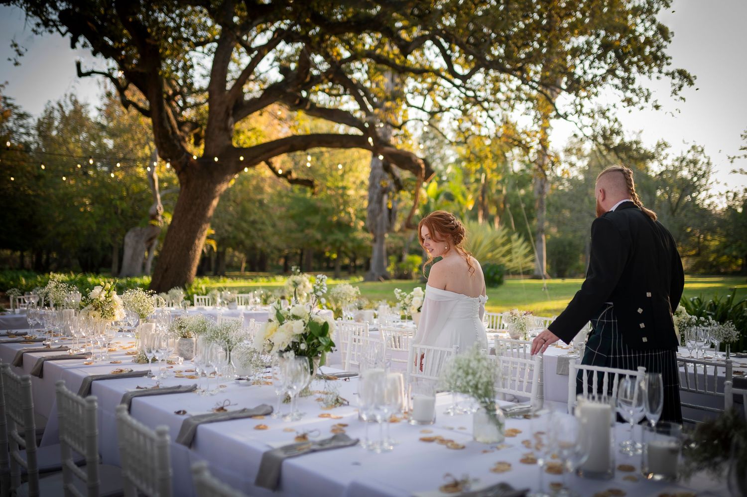 Outdoor wedding reception under shady trees with guests seated at decorated tables on green grass.