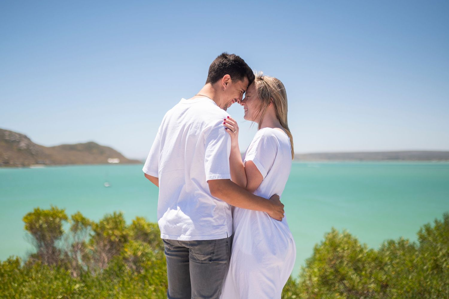 Couple in white shirts embracing and kissing with turquoise ocean and clear blue sky in background.