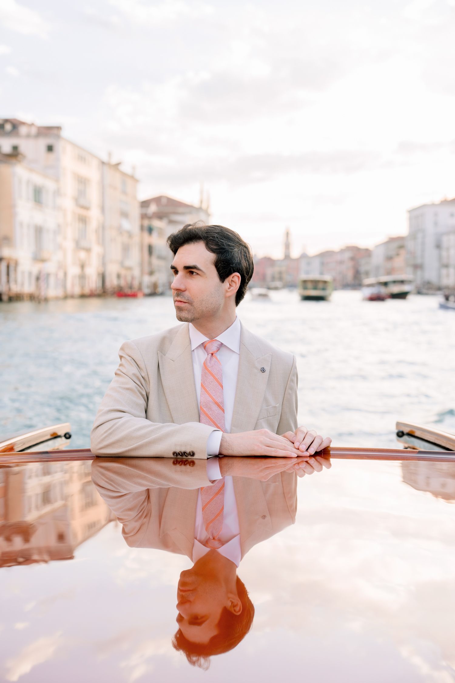 A person in a beige suit and pink tie is photographed reflected in still water against an urban backdrop.