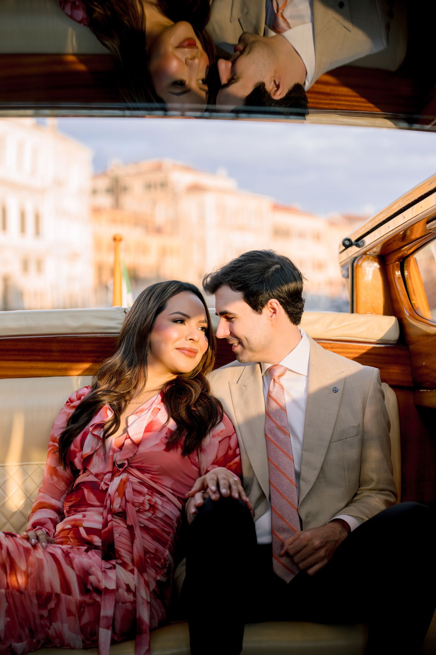 Couple sharing intimate moments on a vintage wooden boat in Venice with historic buildings in background.