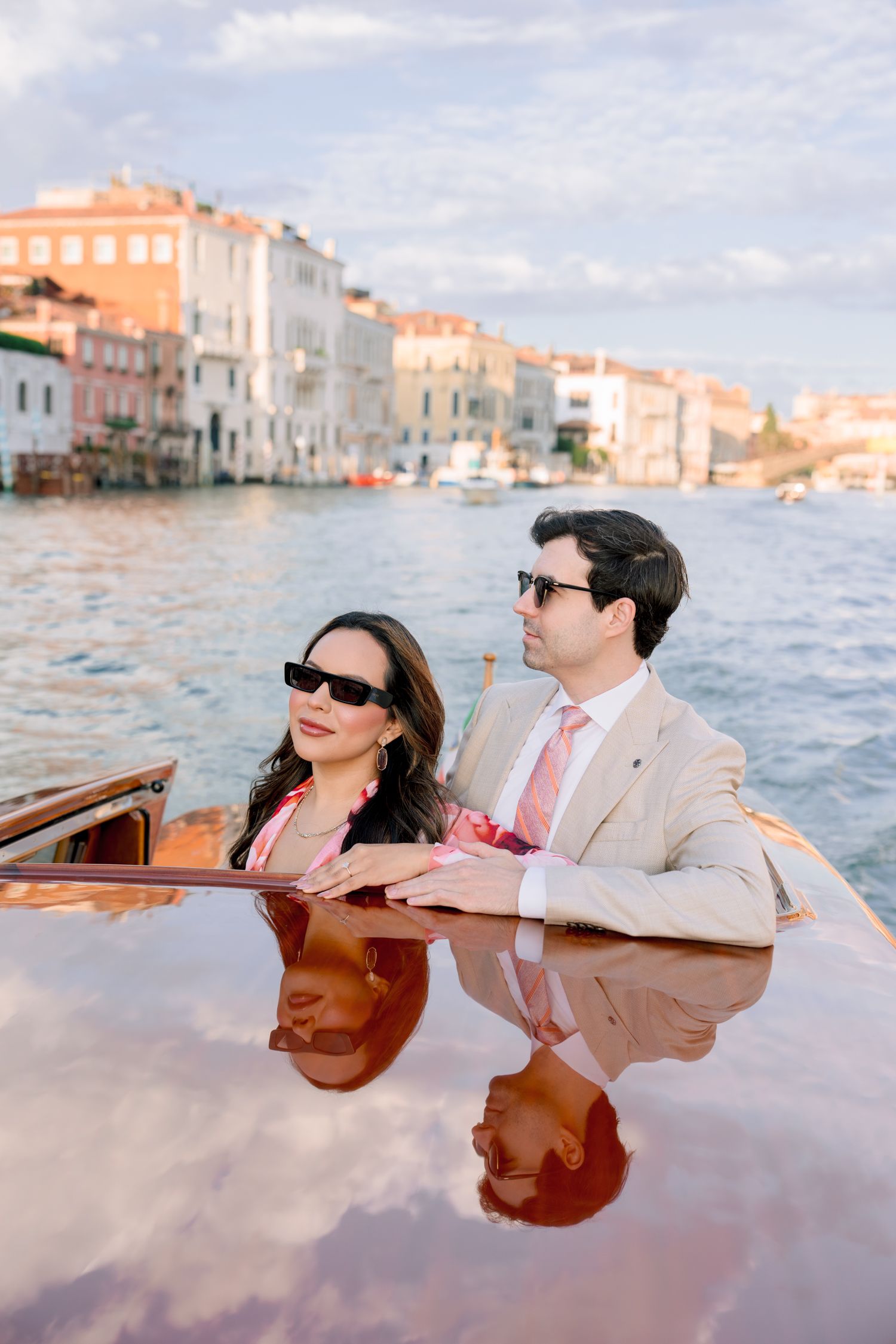 Couple relaxing on a wooden boat in Venice's canals with historic buildings in the background during sunset.