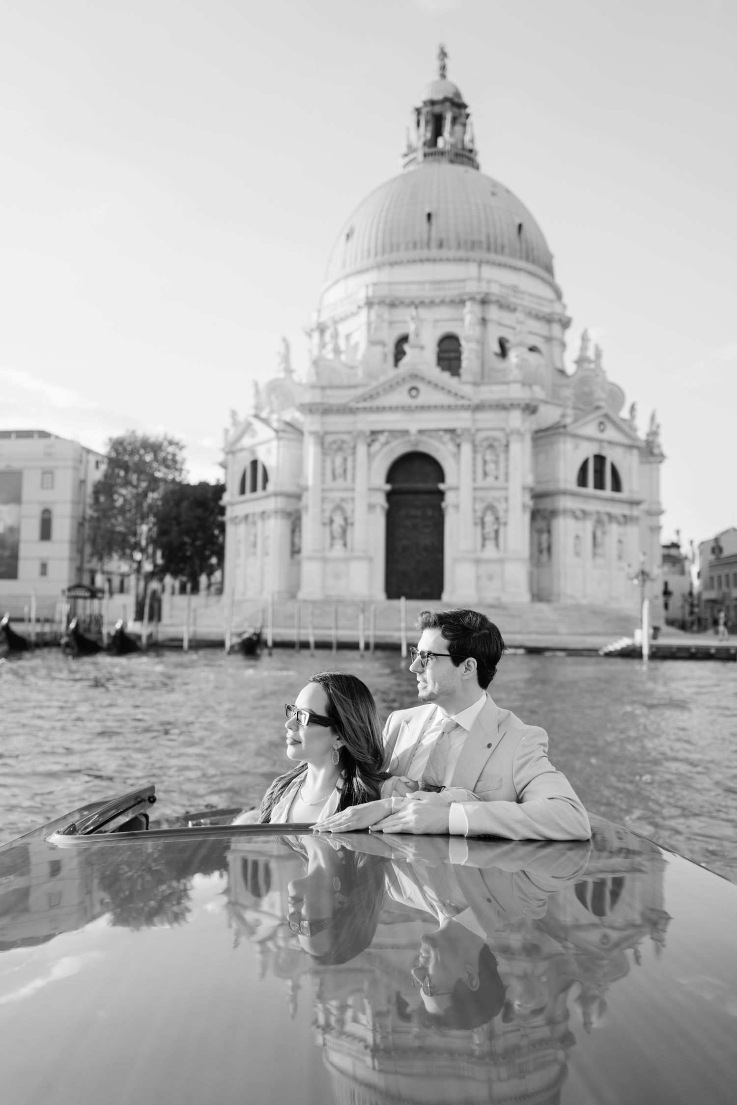 A romantic couple sits in a gondola with the Santa Maria della Salute church reflecting in Venice's calm waters.