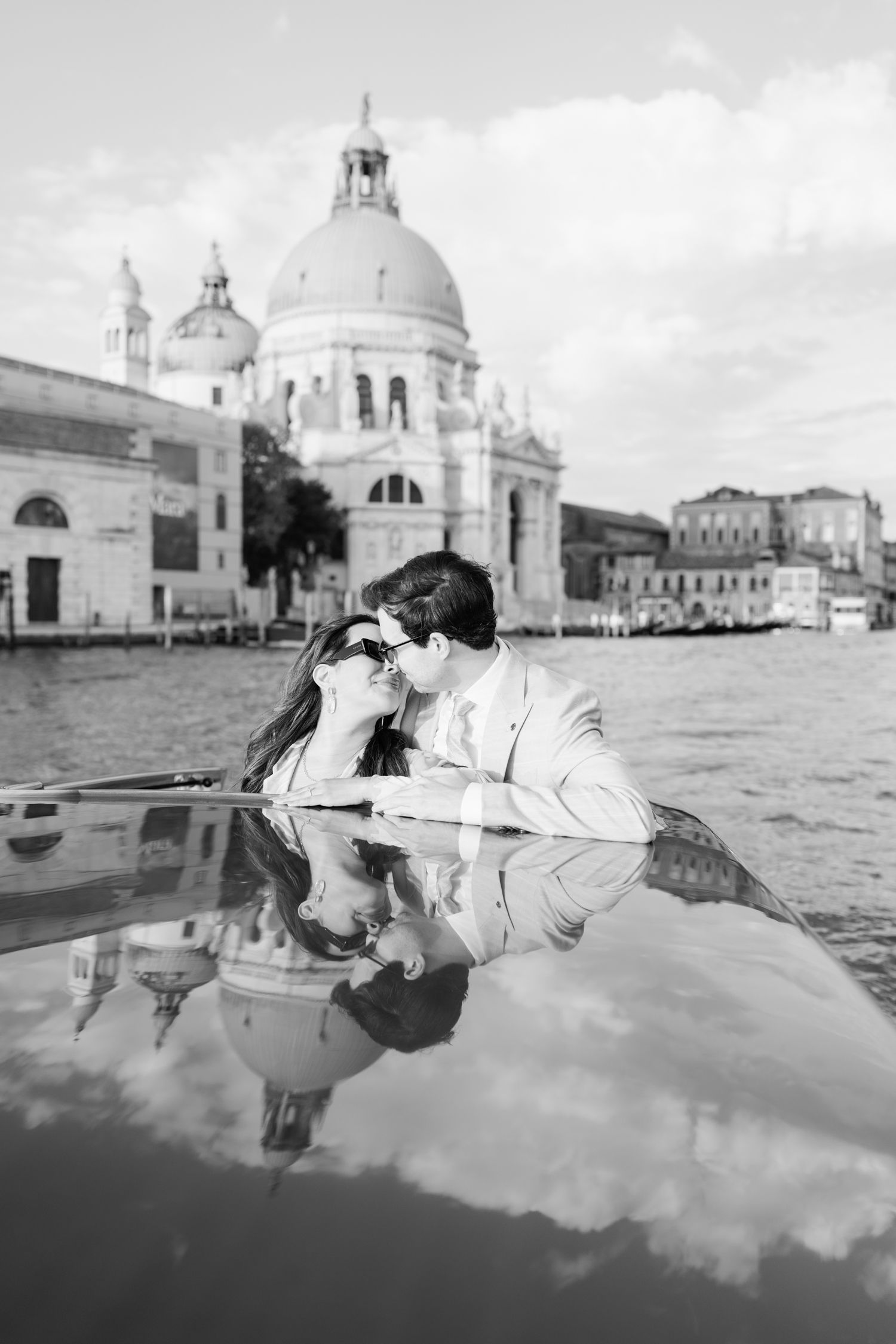 A romantic couple shares a tender moment in a gondola on Venice's Grand Canal with Santa Maria della Salute church in background.