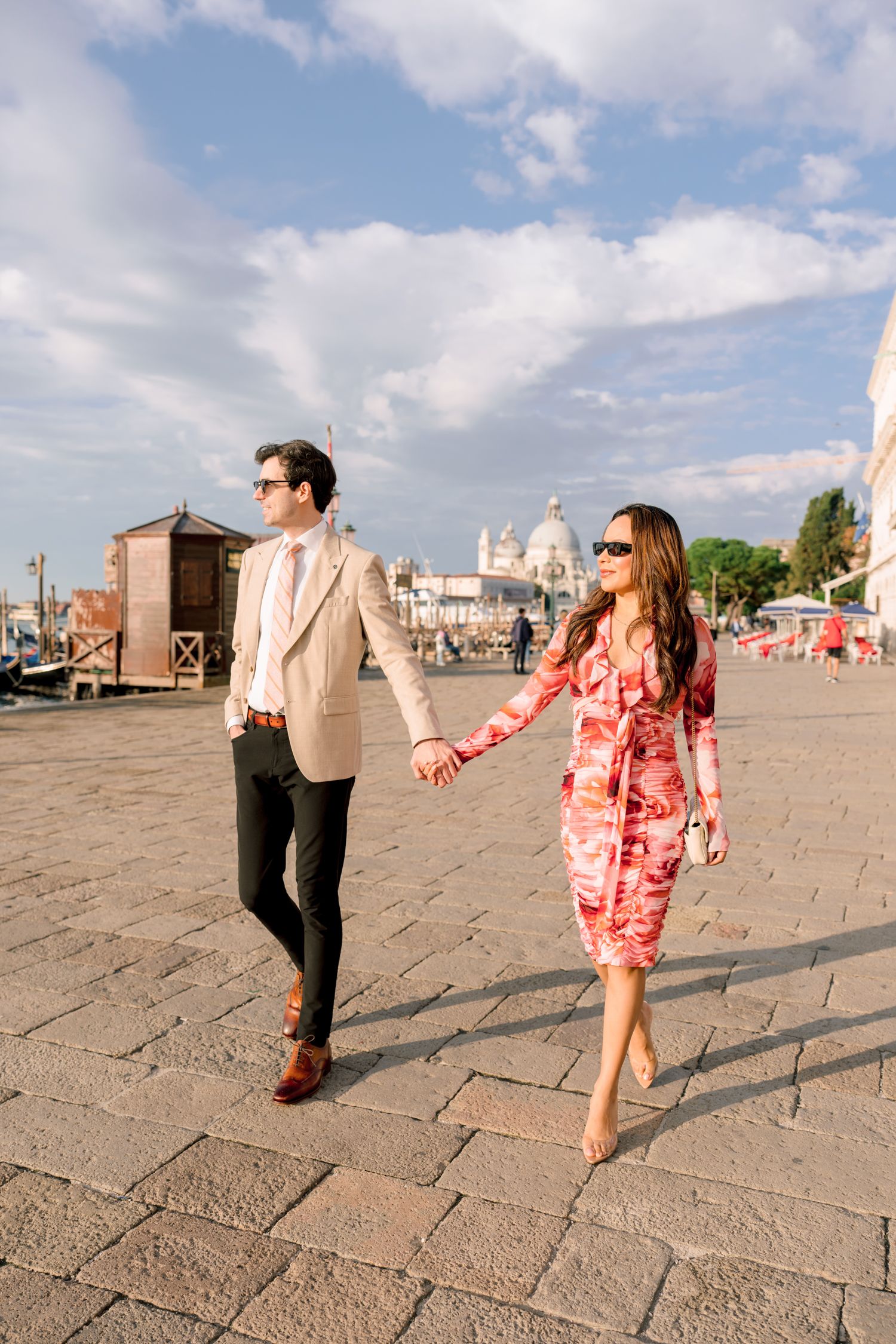 A couple strolls along a cobblestone plaza in Venice with the Basilica di Santa Maria della Salute visible in background.