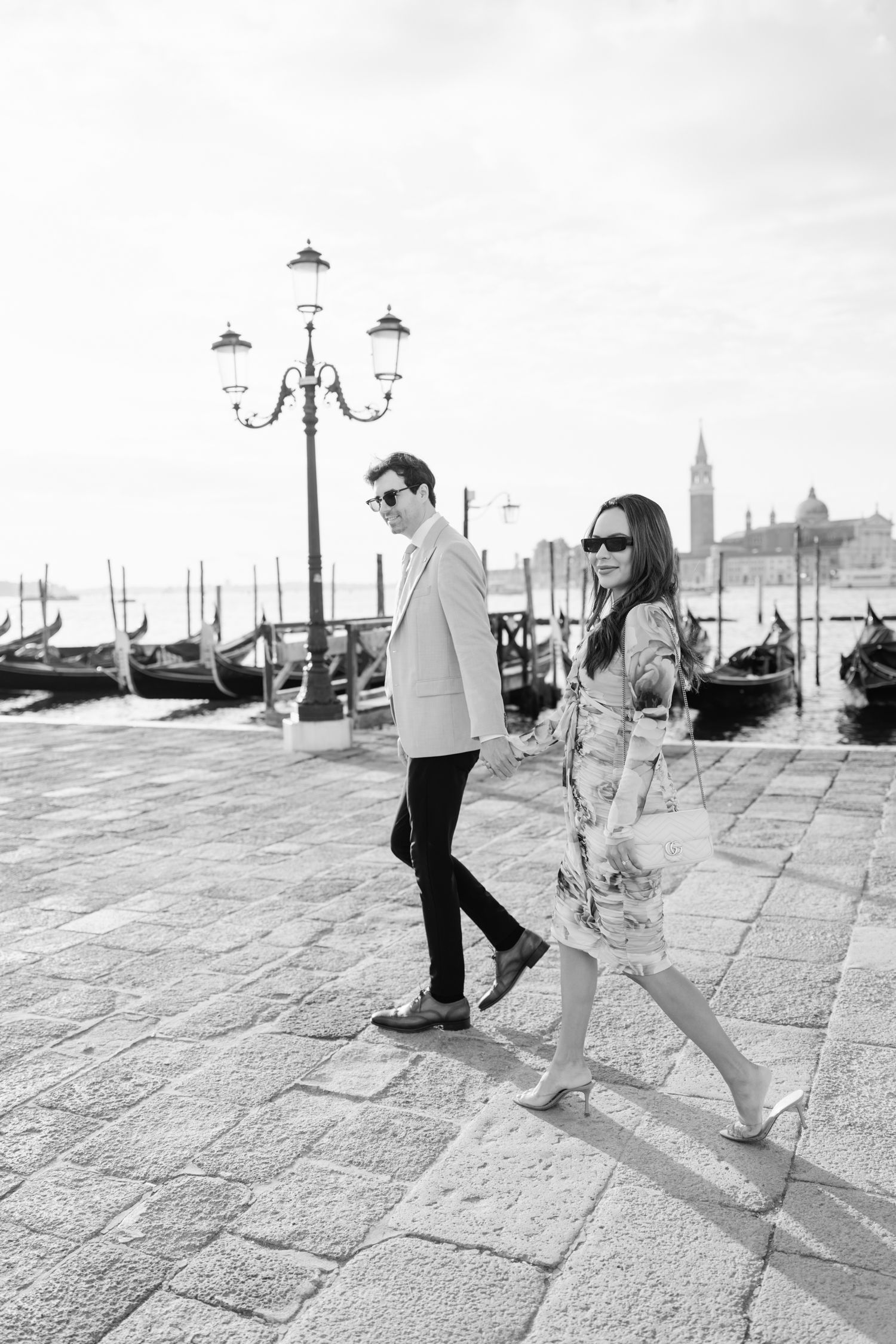 A couple walks along Venice's waterfront with traditional gondolas and ornate street lamps in black and white.